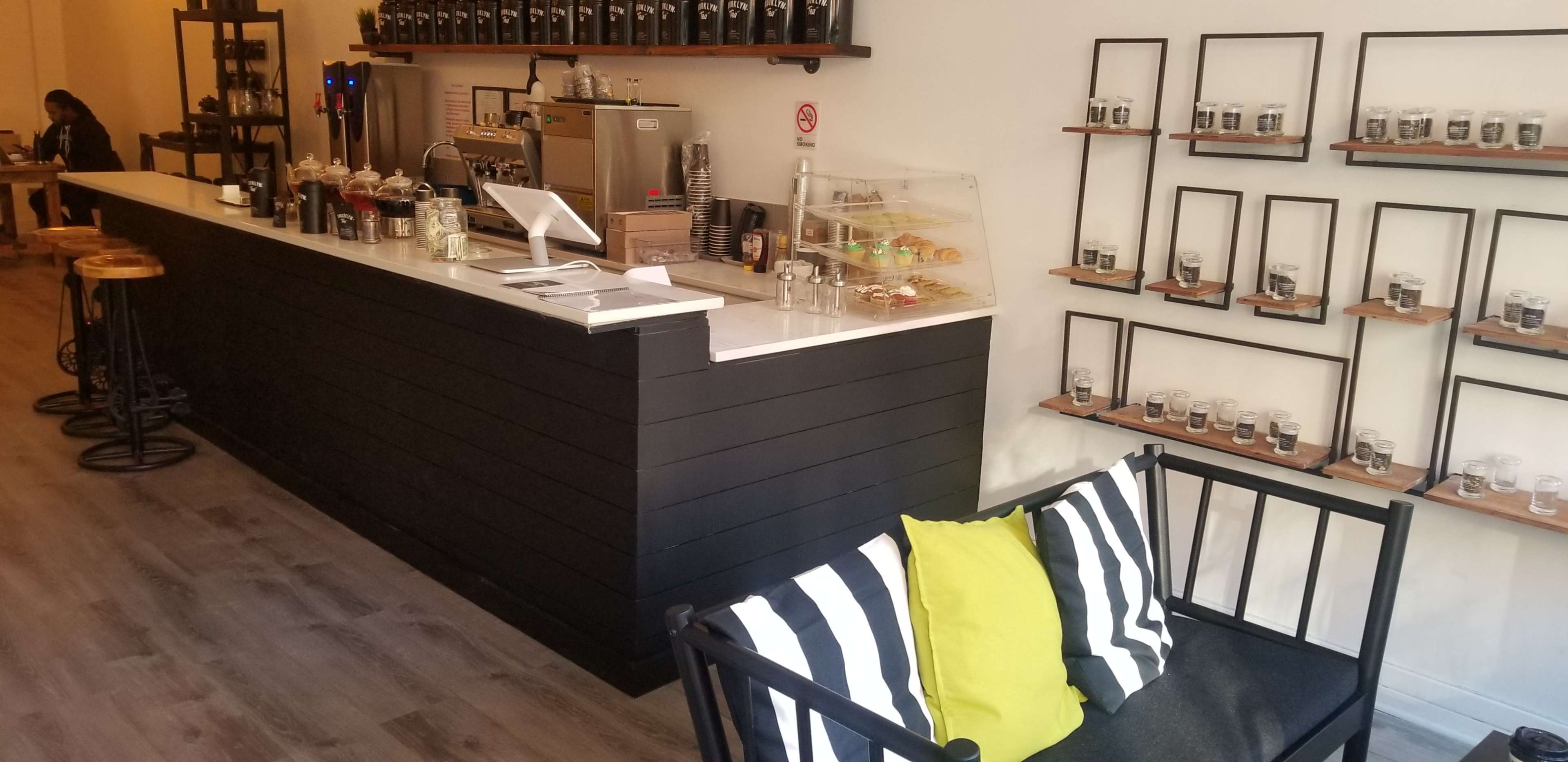 The image shows a modern coffee shop interior featuring a counter with various coffee equipment and a seating area with striped cushions and a bright yellow pillow.