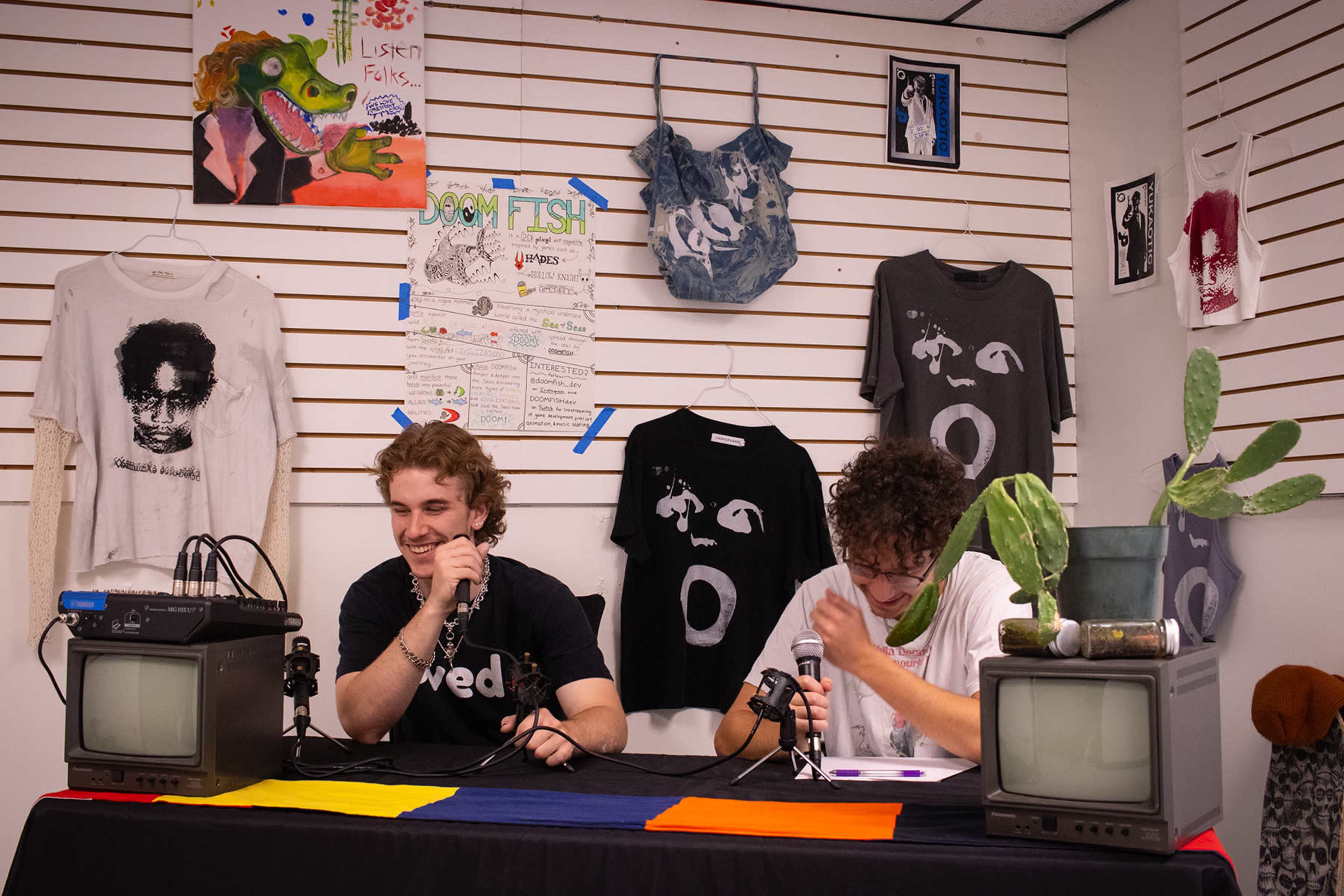 Two young men sit at a table with microphones in front of them, surrounded by artwork and hanging shirts on a wall.