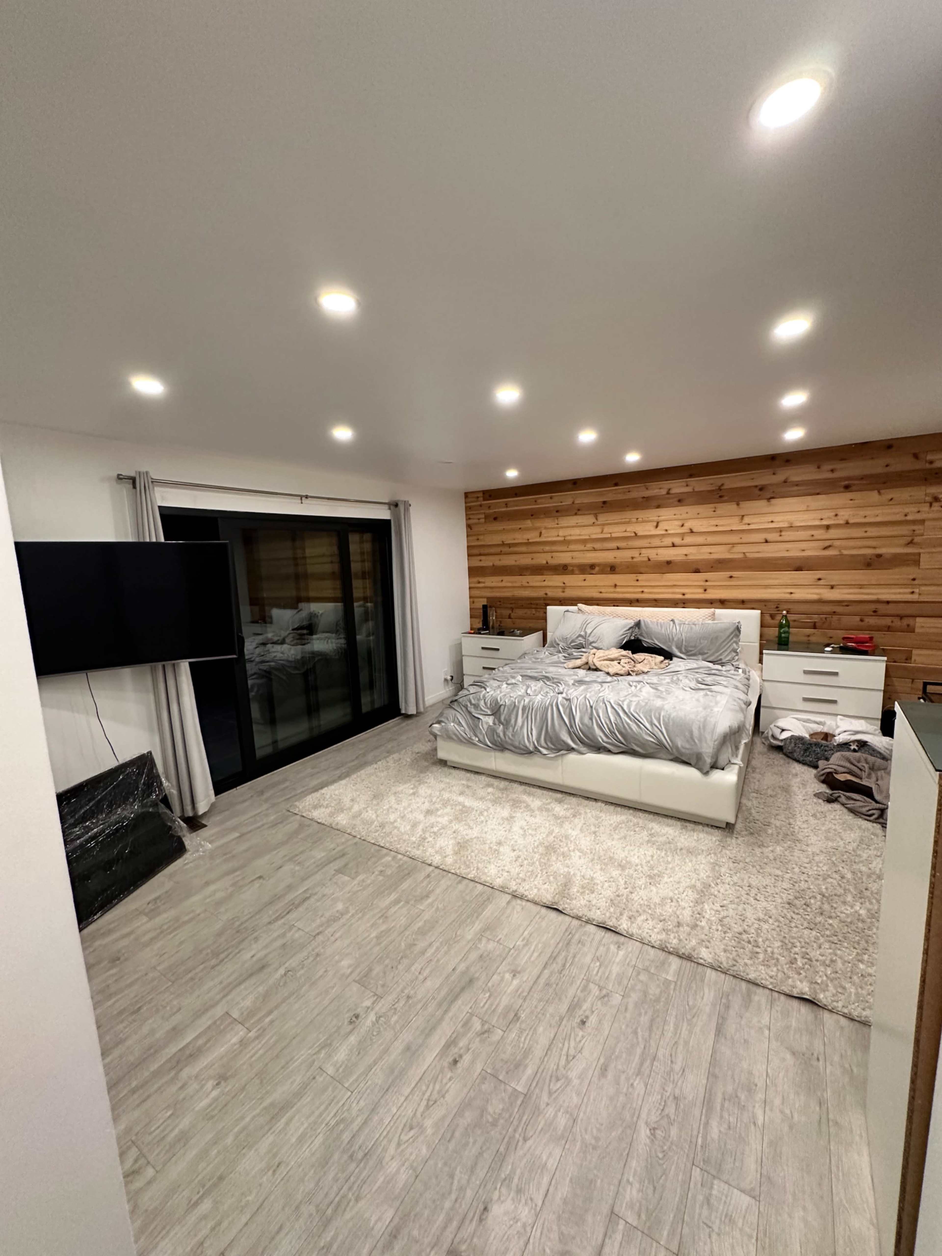The image shows a modern bedroom with a large bed covered in gray bedding, a wooden accent wall, and a sliding glass door leading to an outside area.