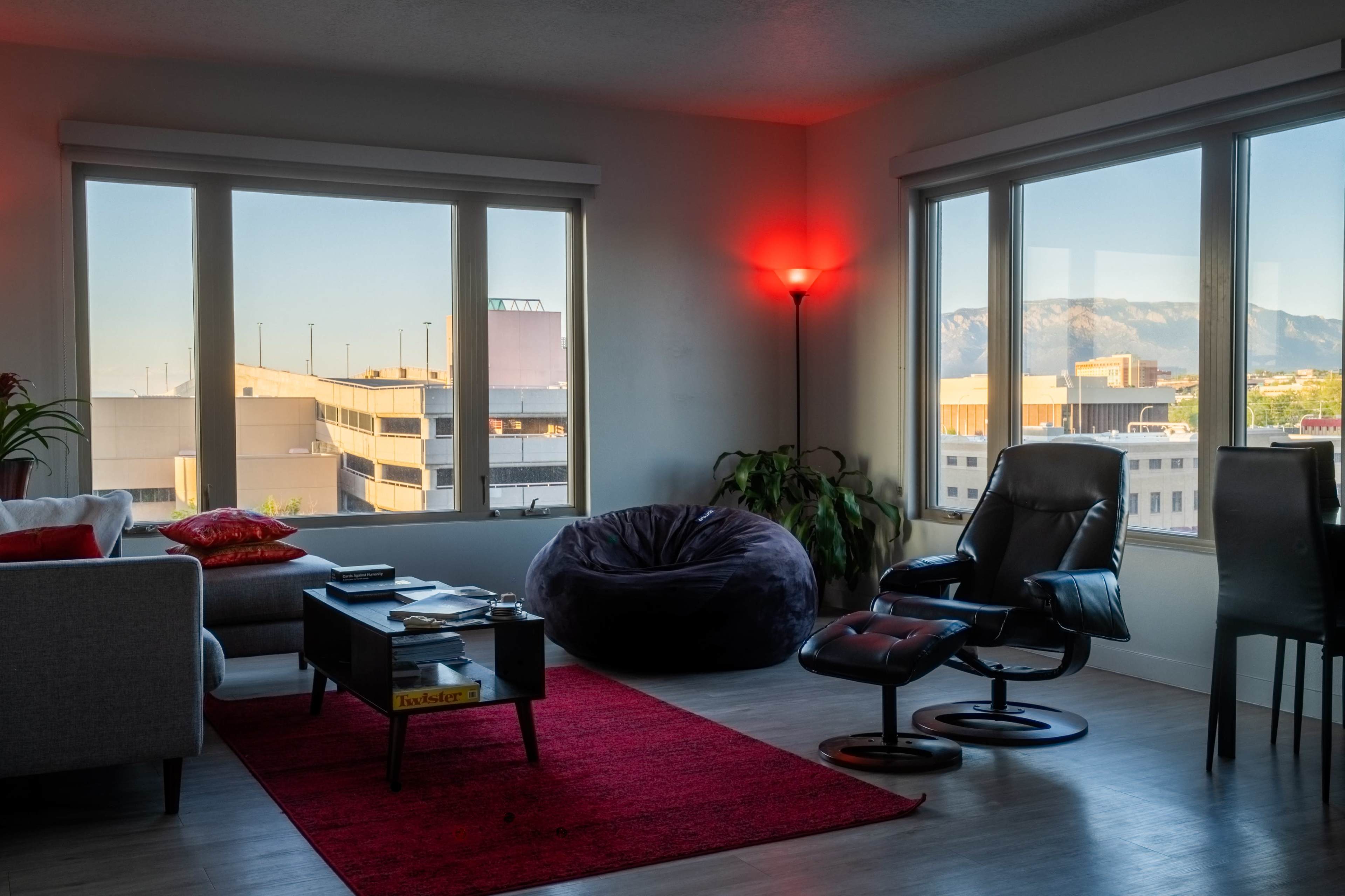 A modern living room features large windows with a mountain view, a red area rug, a bean bag chair, and a comfortable recliner beside a small coffee table.
