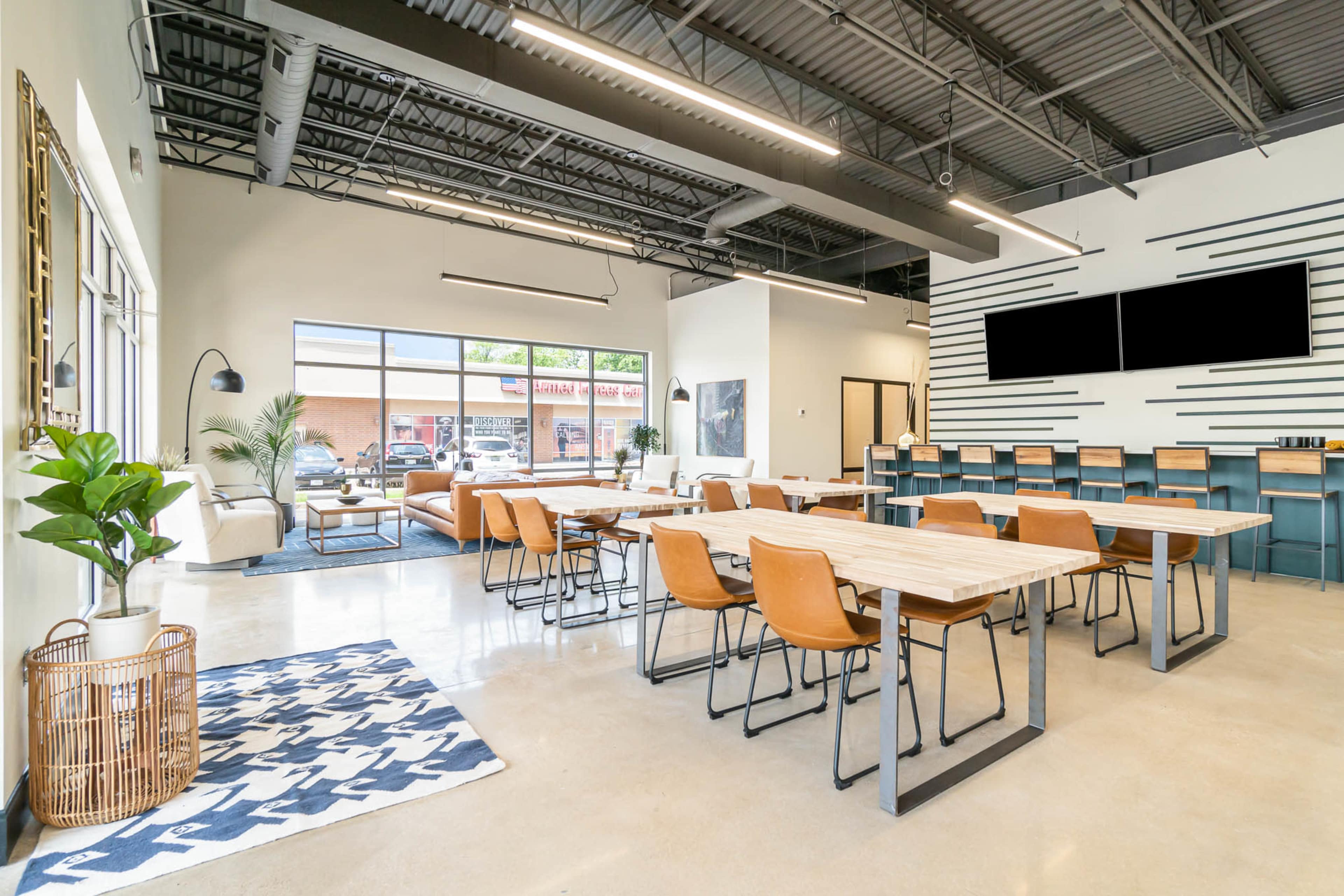 A spacious, modern work area features multiple tables and chairs, large windows letting in natural light, and a seating area with a plant.