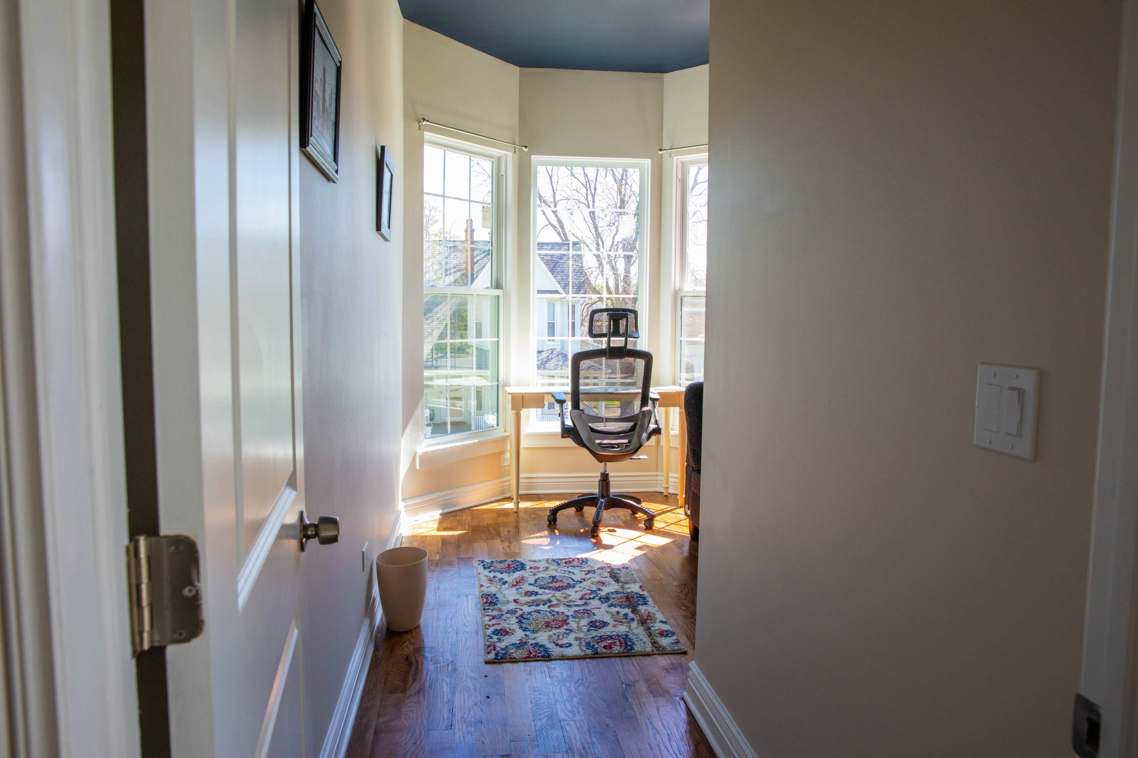 A narrow hallway leads to a bright corner home office with a chair and desk near large windows.