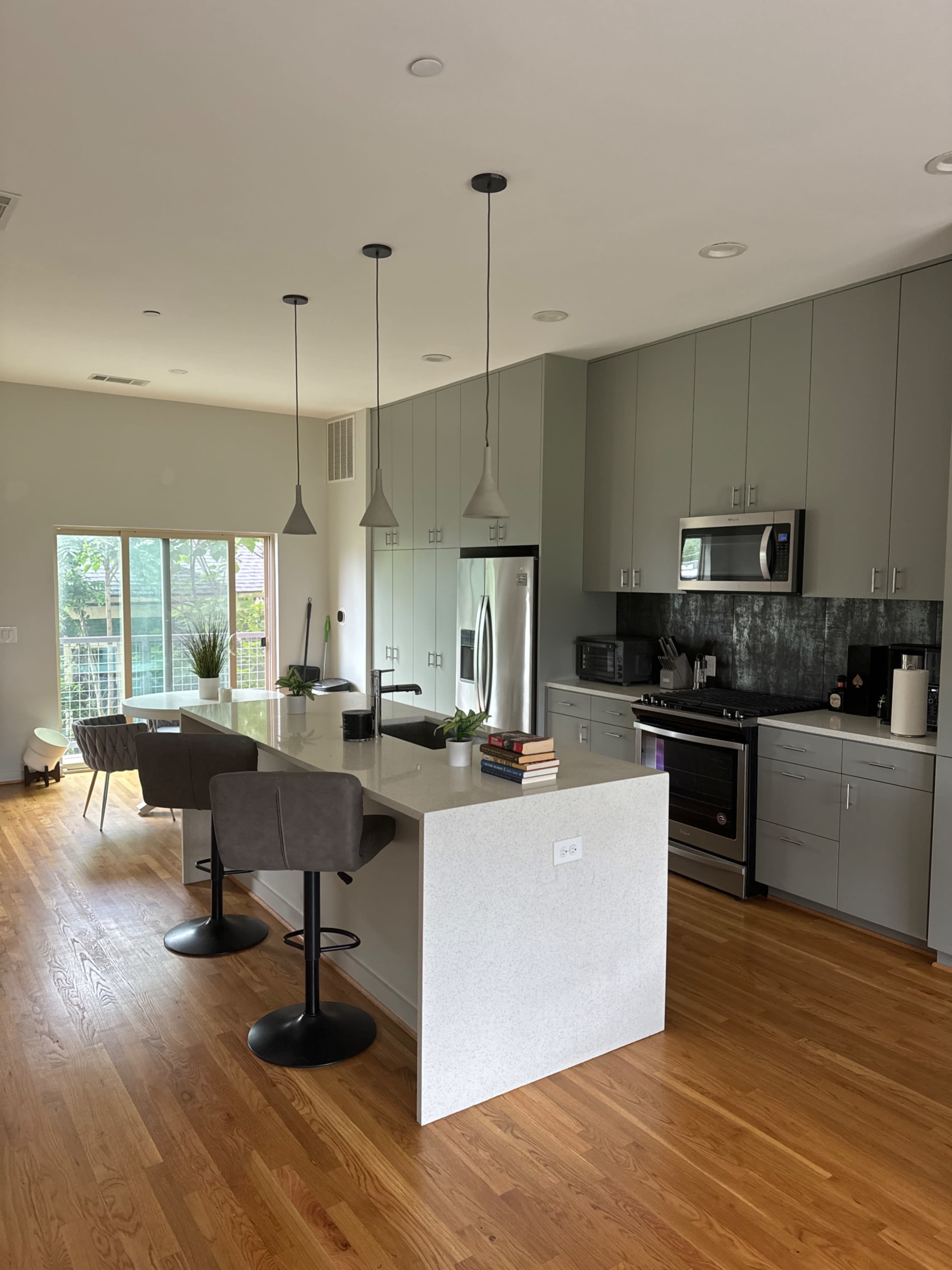 A modern kitchen with an island featuring bar stools, gray cabinetry, stainless steel appliances, and large windows allowing natural light.