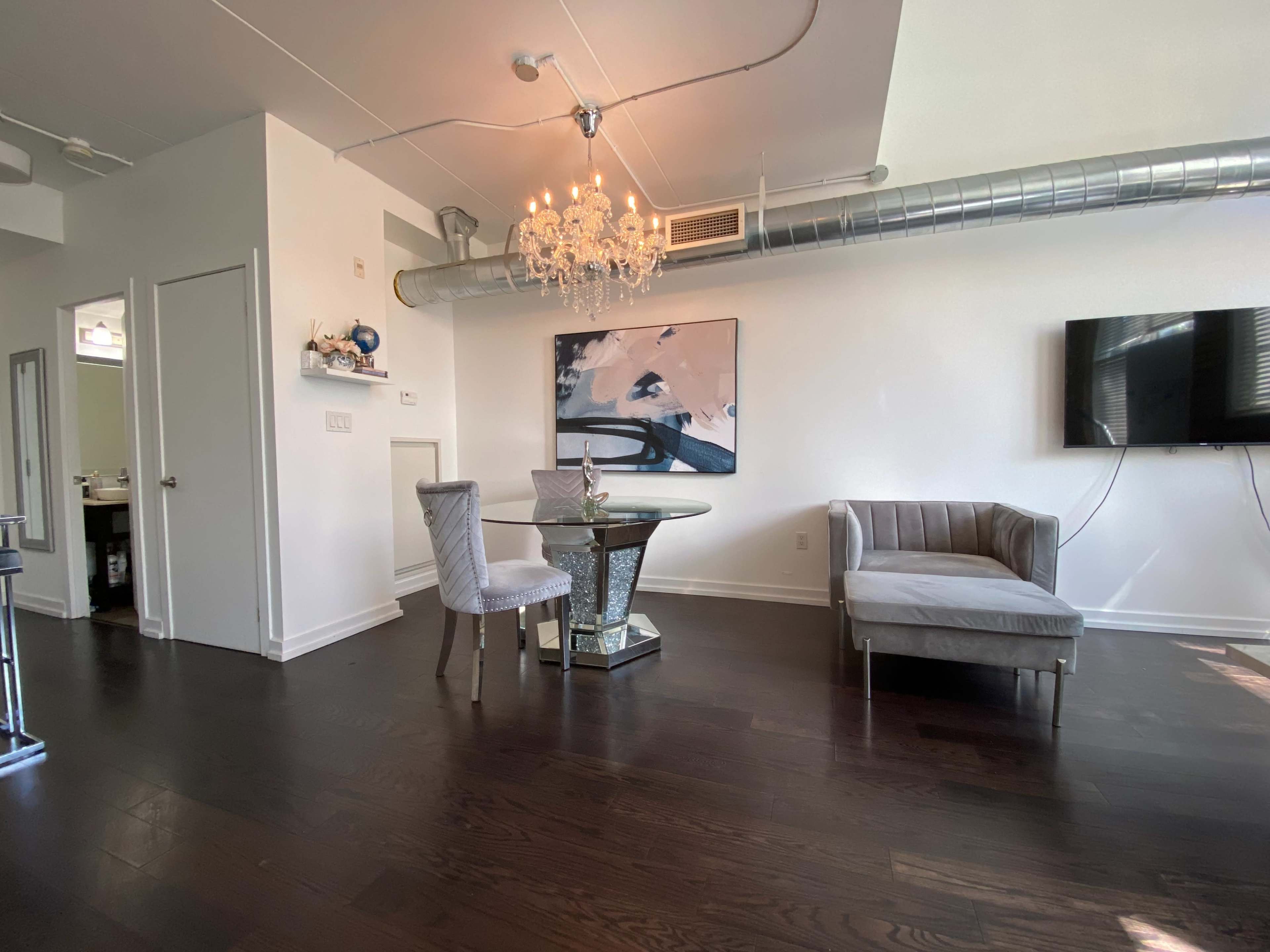 A modern living space featuring a round dining table with two chairs, a small sofa, a wall-mounted TV, and a chandelier.