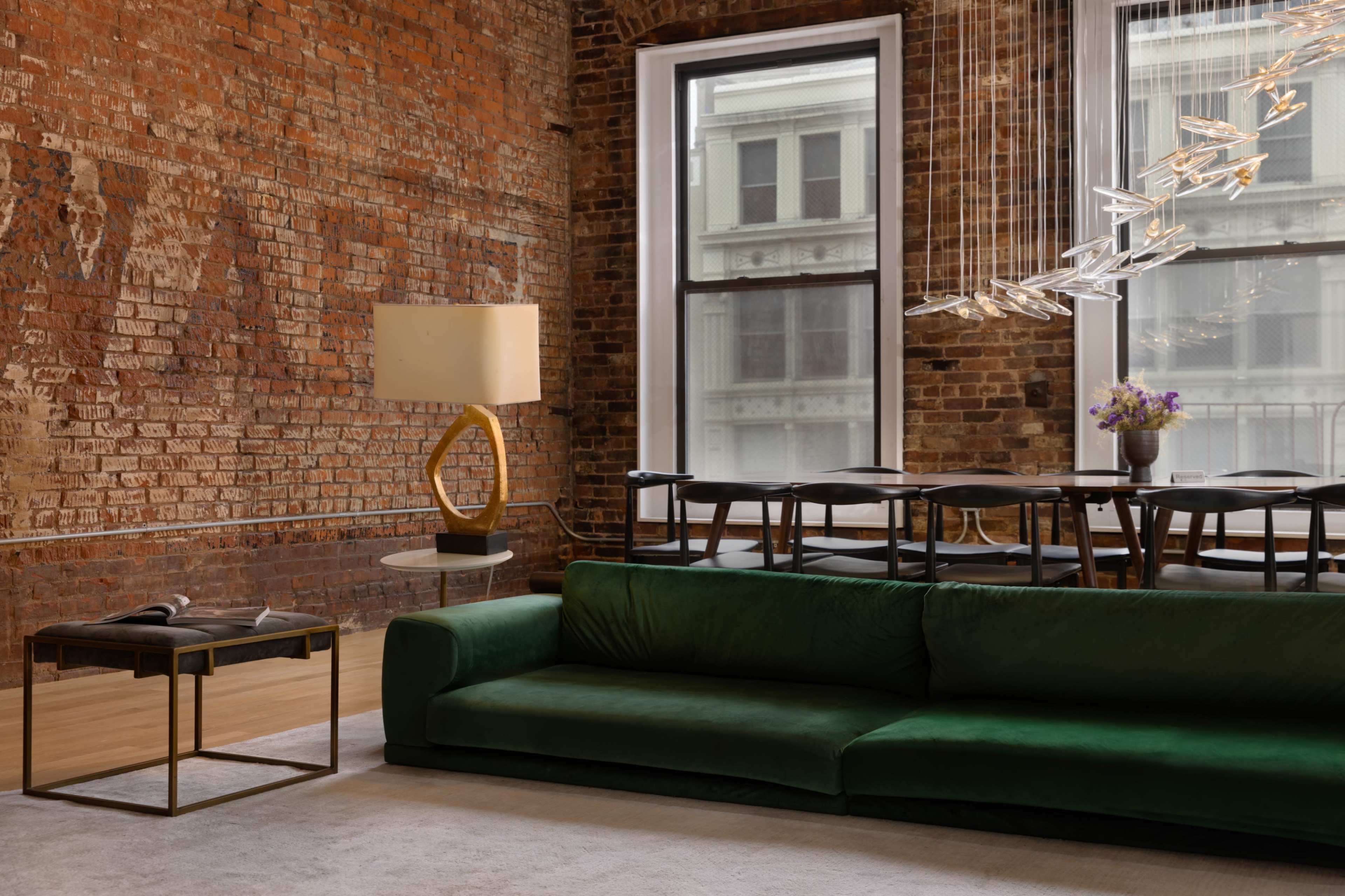 A green sofa sits in front of a rugged brick wall, accompanied by a round coffee table, a modern floor lamp, and a dining table with chairs beneath stylish light fixtures.