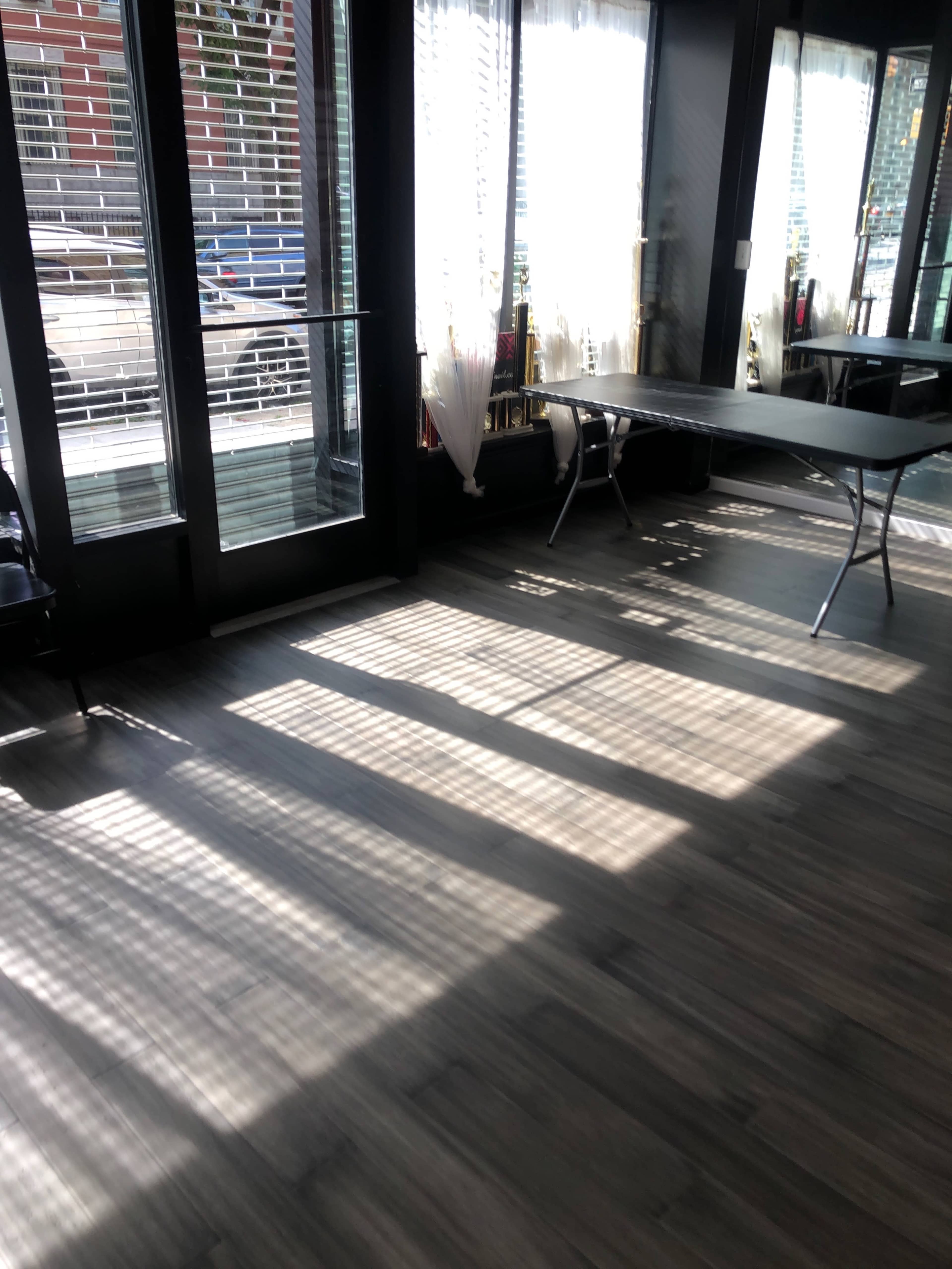 The image shows a room with large windows casting striped shadows on a wooden floor, accompanied by a couple of folding tables.