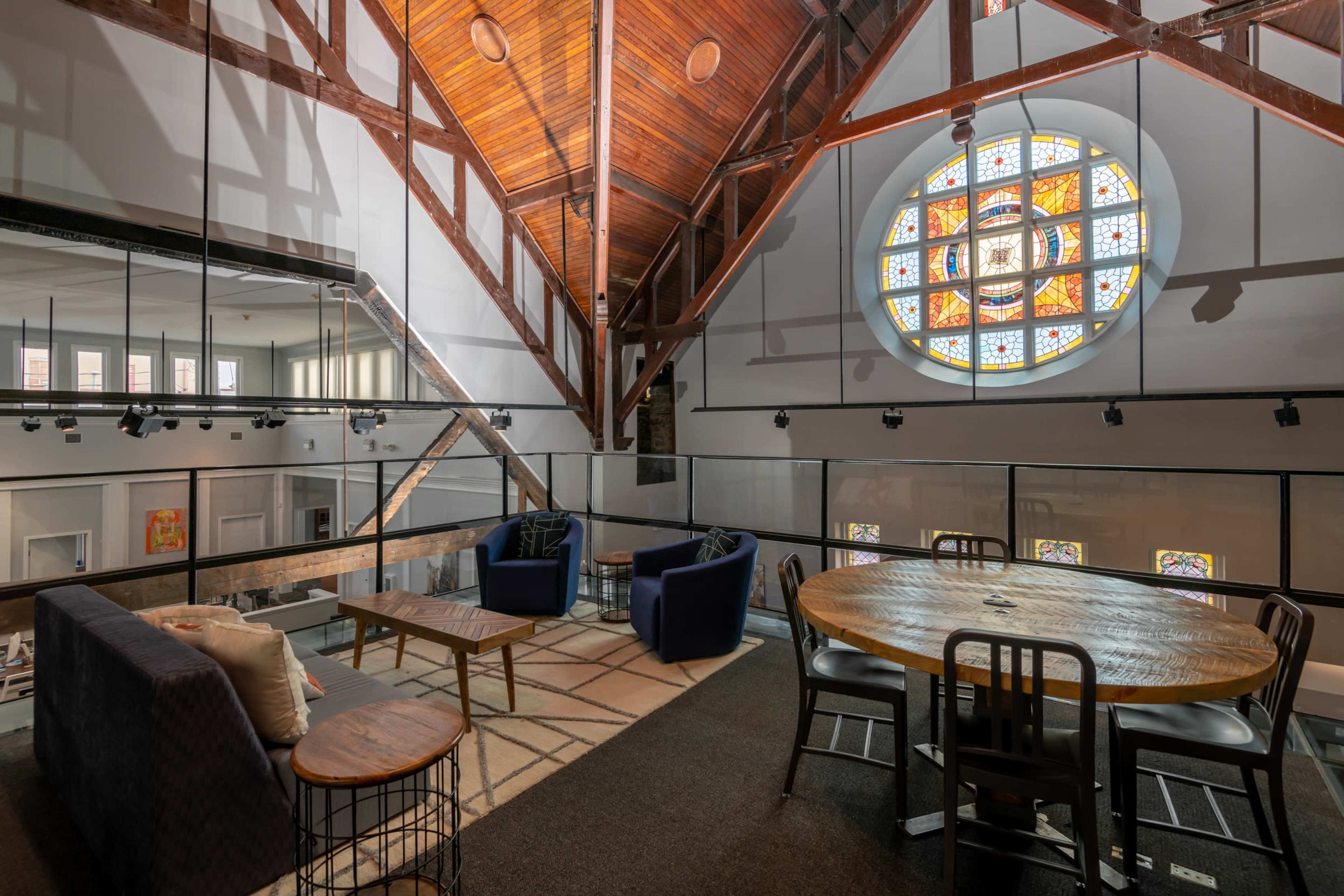 A spacious room with high wooden ceilings, featuring a round dining table and chairs in the foreground, and a large stained glass window in the background.