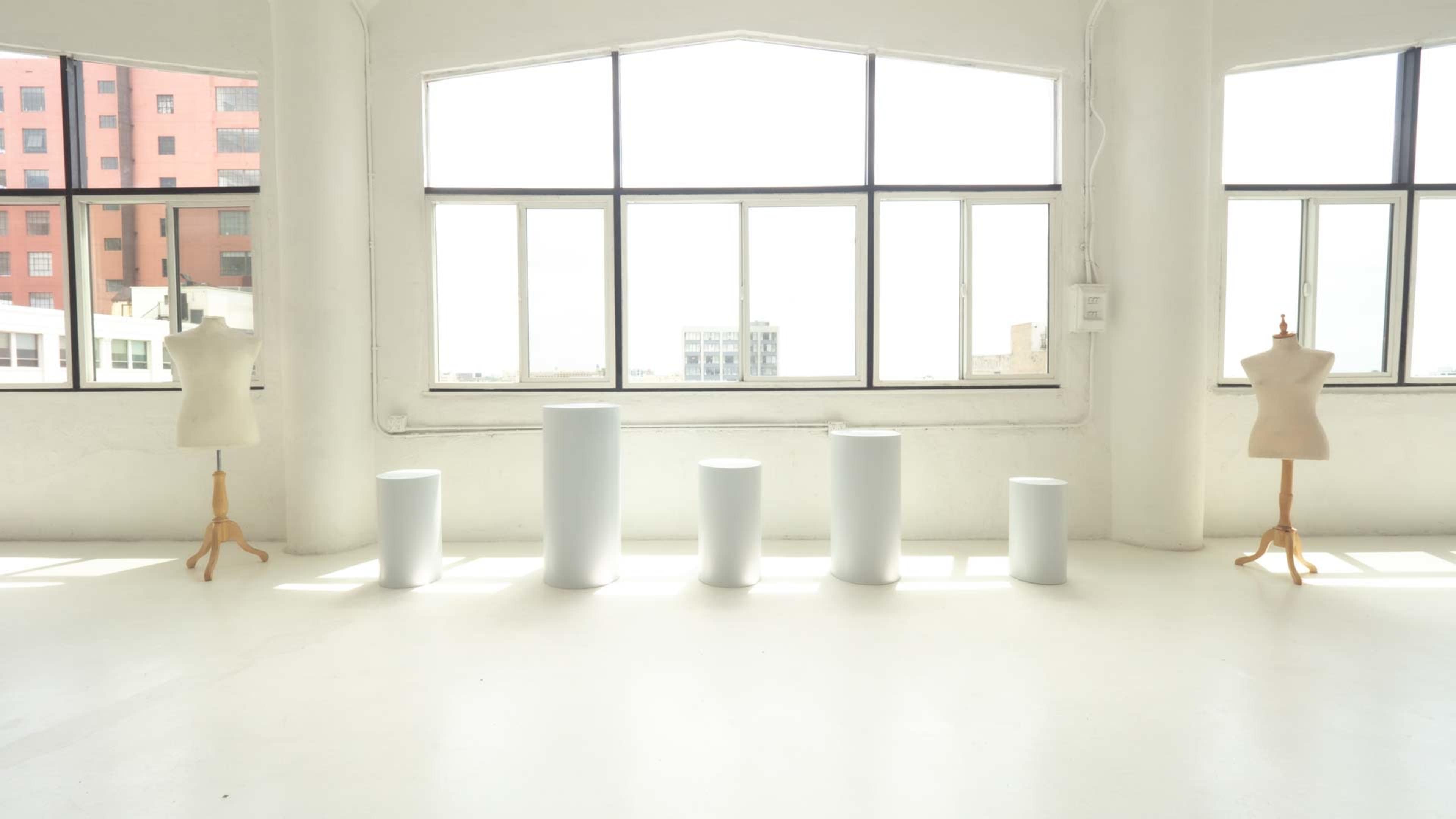A bright, white studio space with five white cylindrical pedestals arranged in a row and two mannequin forms positioned nearby.