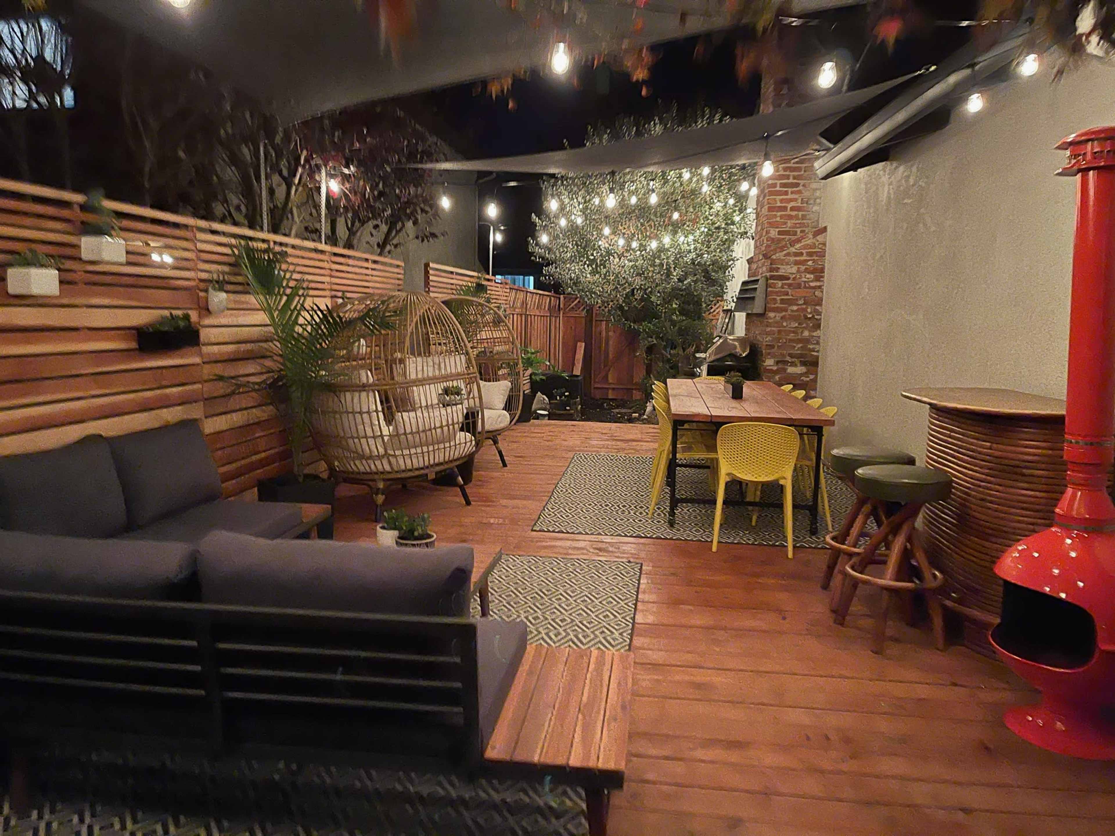A modern outdoor patio with wooden decking, seating areas, a dining table, and string lights overhead.