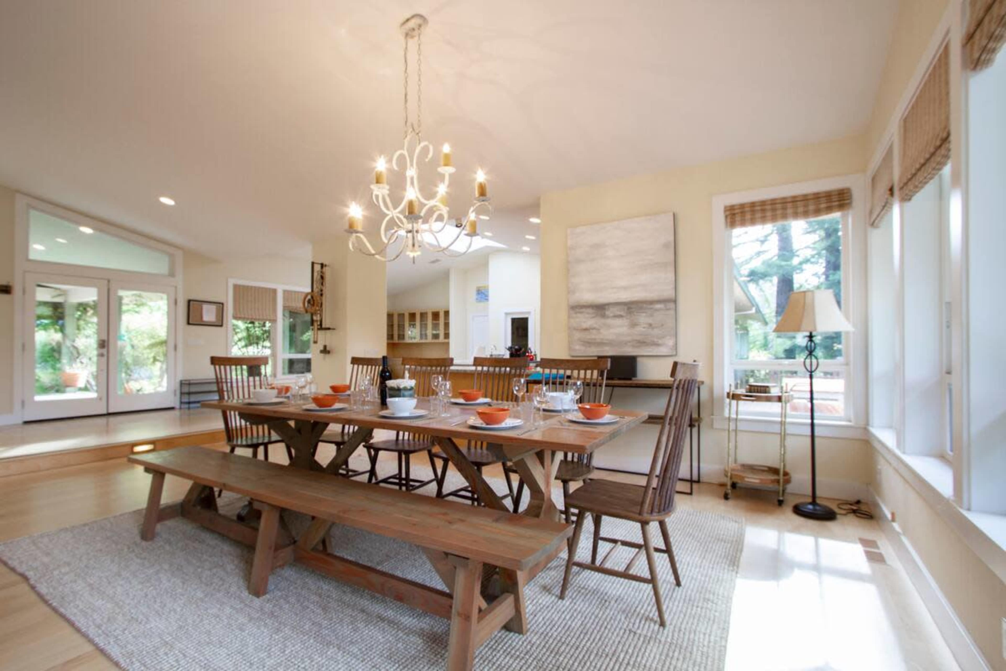 A spacious dining room features a large wooden table set for a meal, surrounded by simple wooden chairs, with a chandelier overhead and windows letting in natural light.