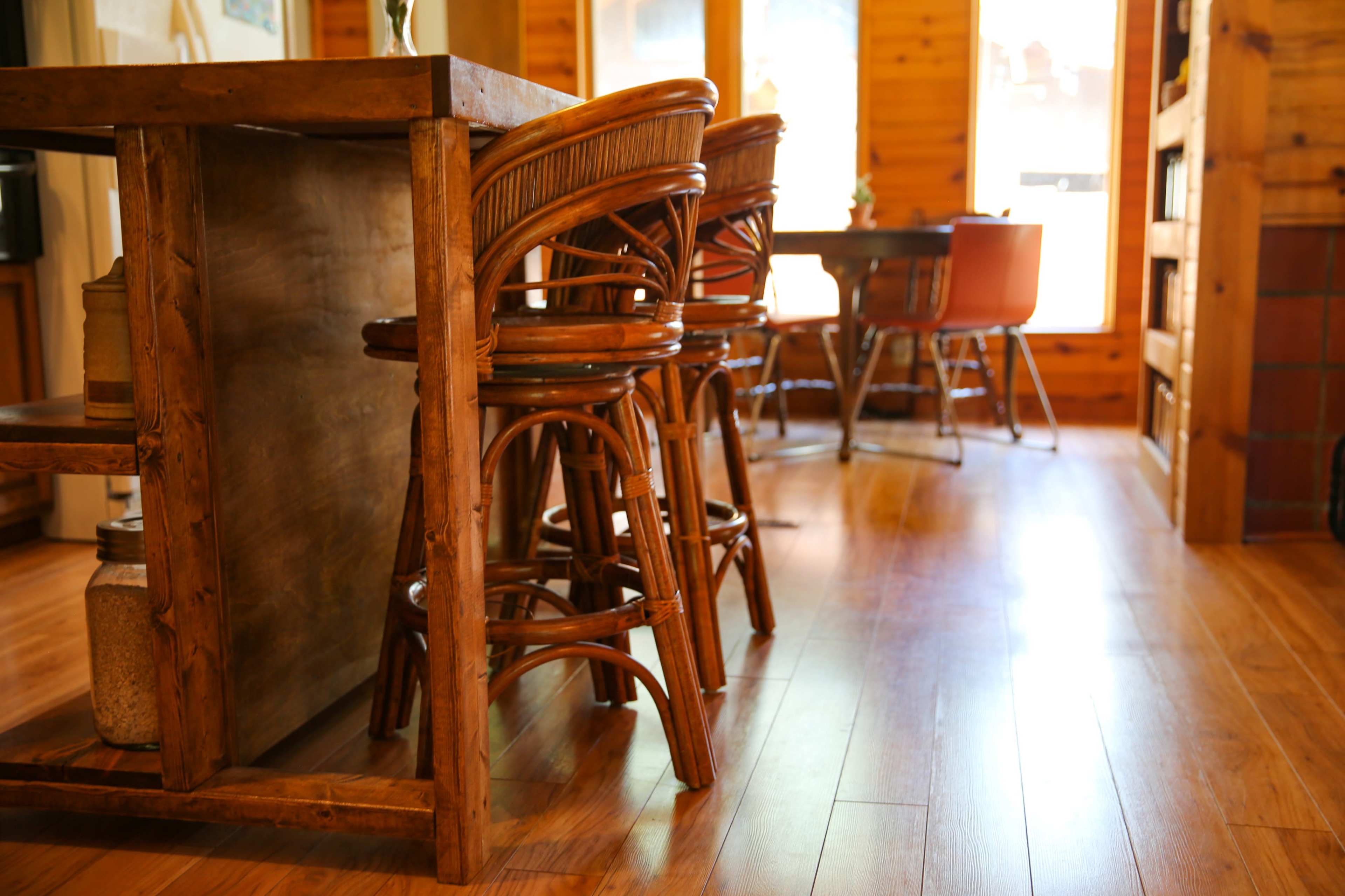 A wooden kitchen area with bar stools beside a counter and a dining table in the background.
