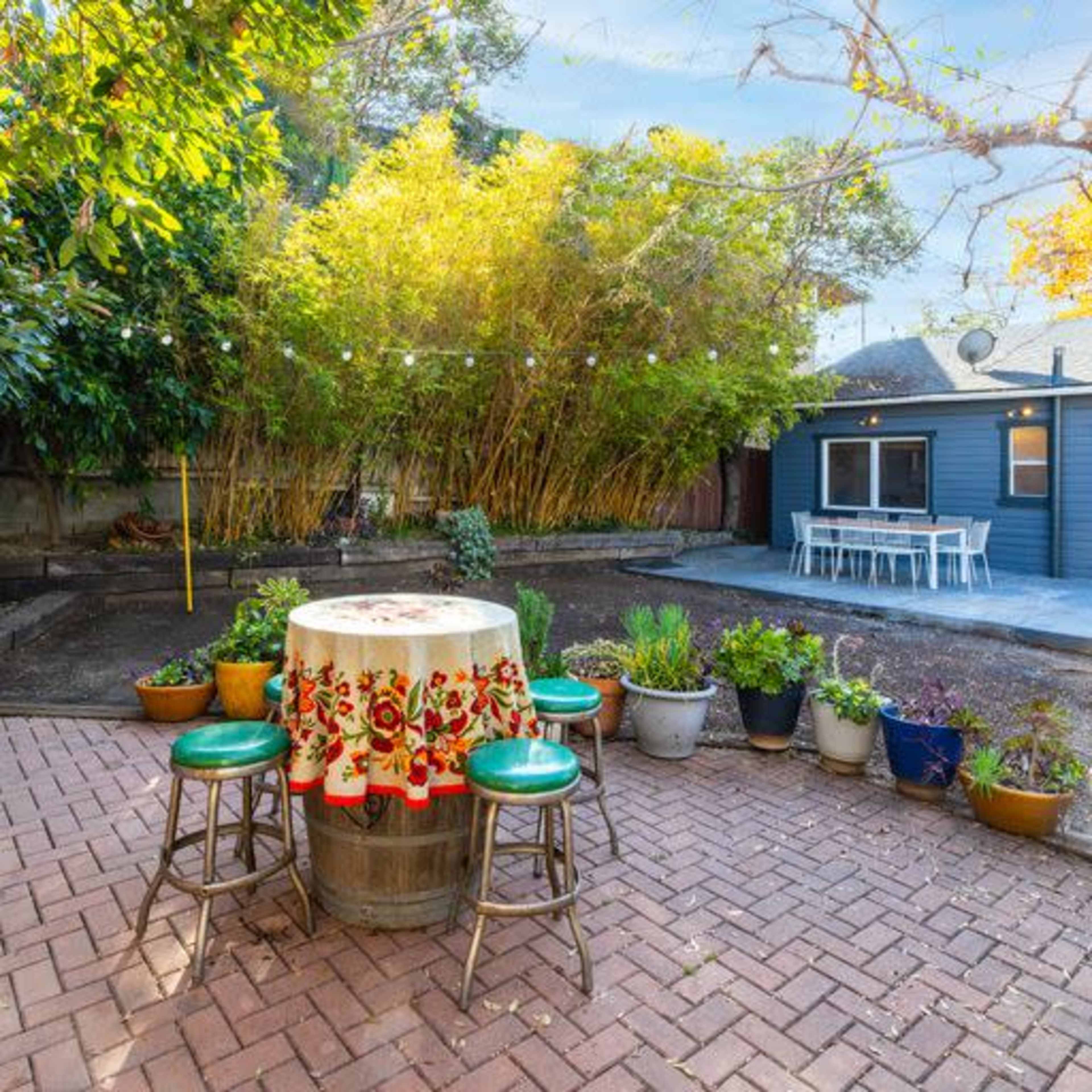 A backyard with a brick patio, a round table with decorative fabric, potted plants, and a blue building in the background.