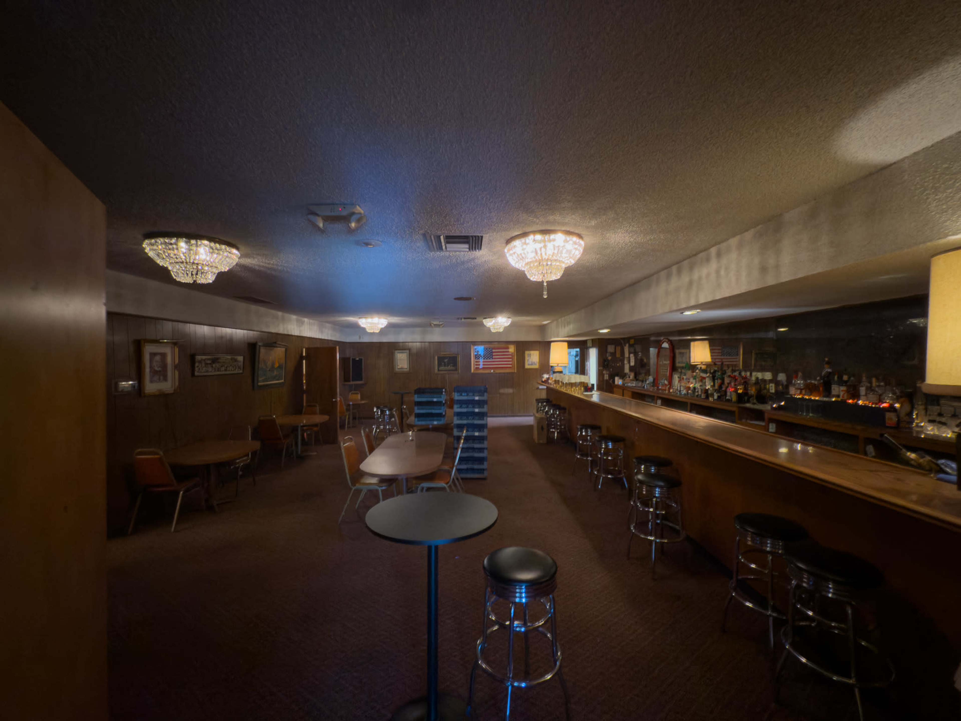 The image shows a dimly lit bar with a long wooden counter, scattered tables, and plush seating arranged in a cozy interior space.