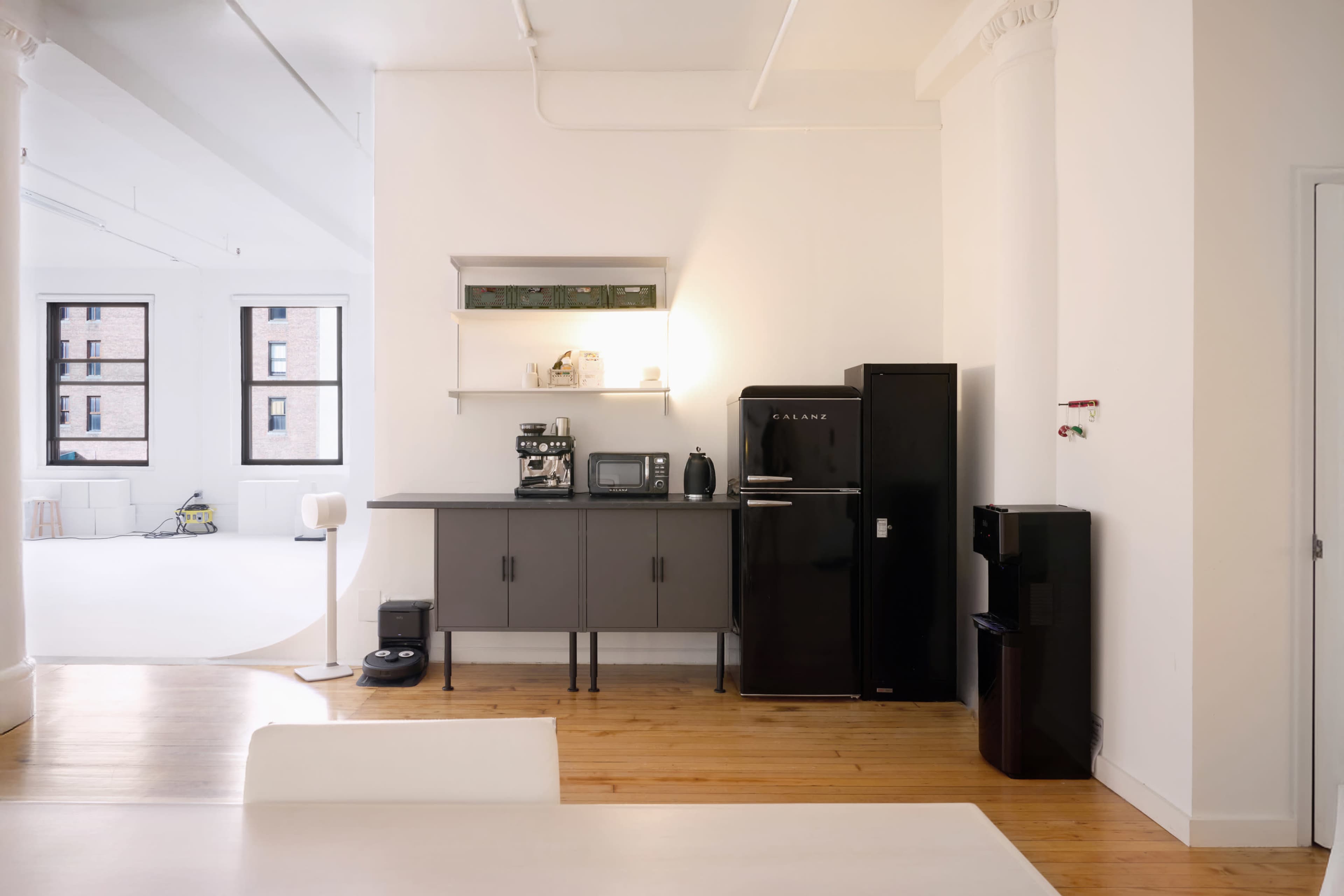 The image shows a modern kitchen area with a black refrigerator, a microwave, a coffee maker, and a water cooler against a white wall.