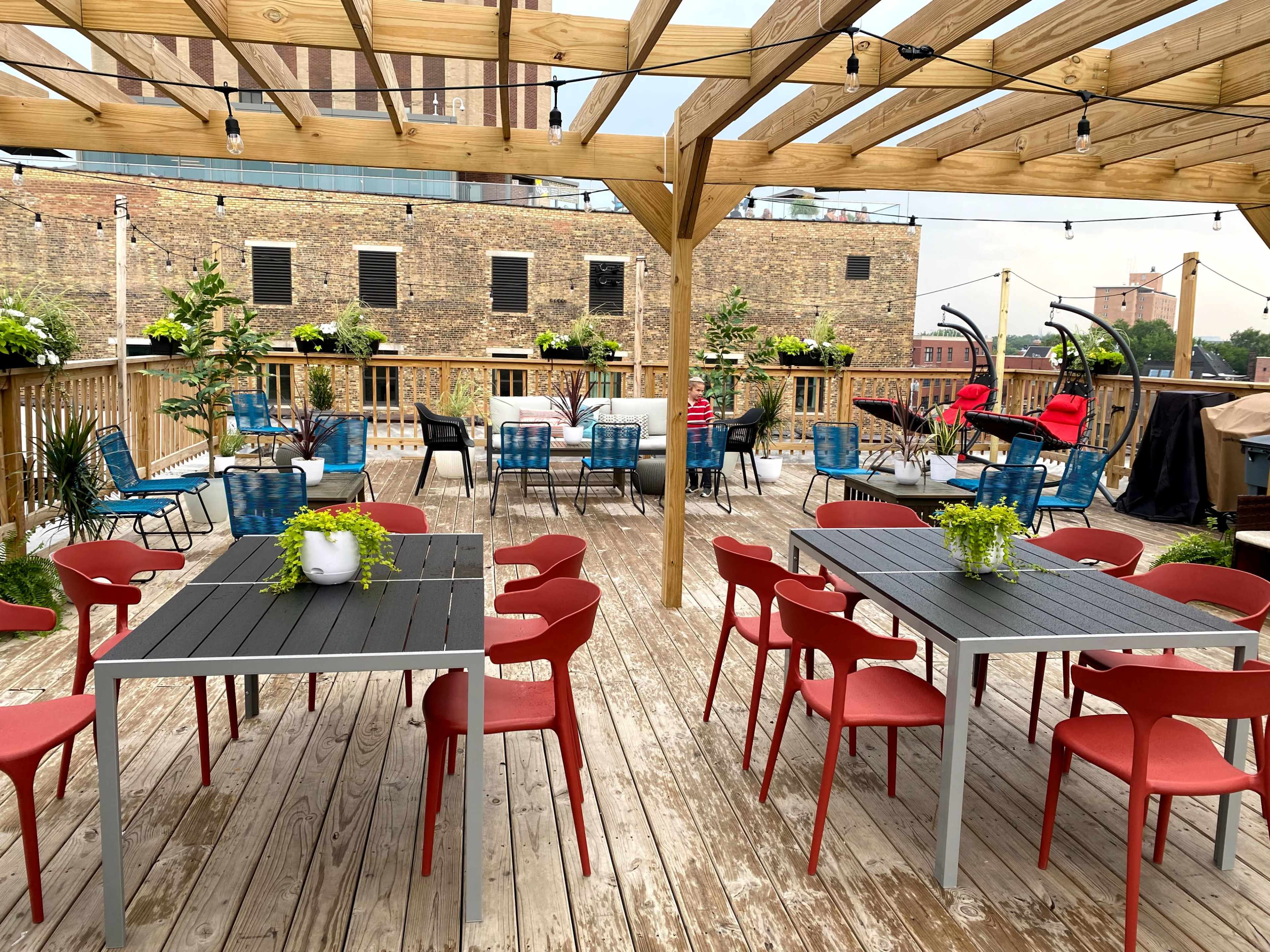A rooftop terrace features several tables and chairs, with green plants arranged around the wooden deck and a view of nearby buildings.