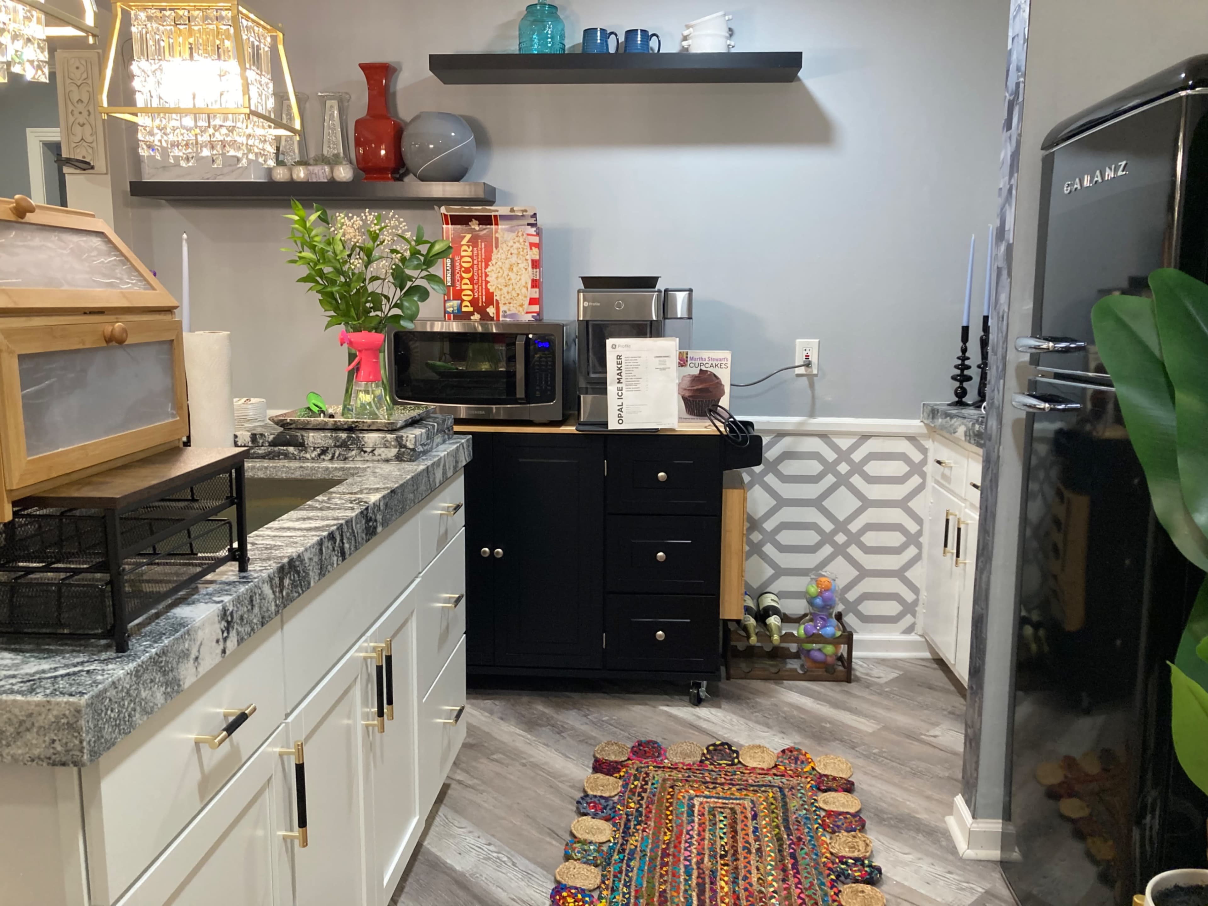 A kitchen area with a black microwave, a small black cabinet, and decorative items on shelves, along with a colorful rug on the floor.