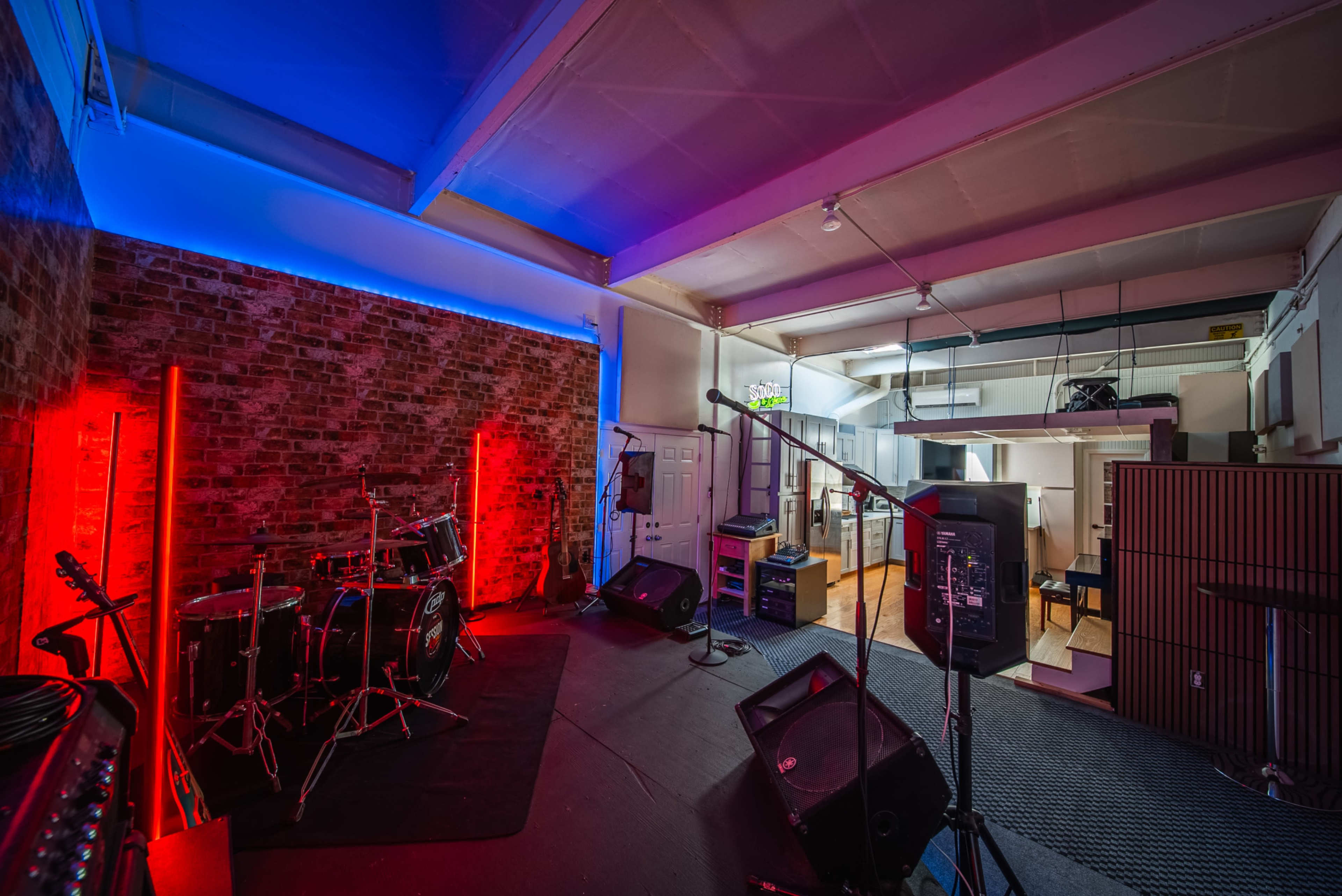 The image shows a music rehearsal space with a brick wall, stage lights in red and blue, a drum set, and various audio equipment.