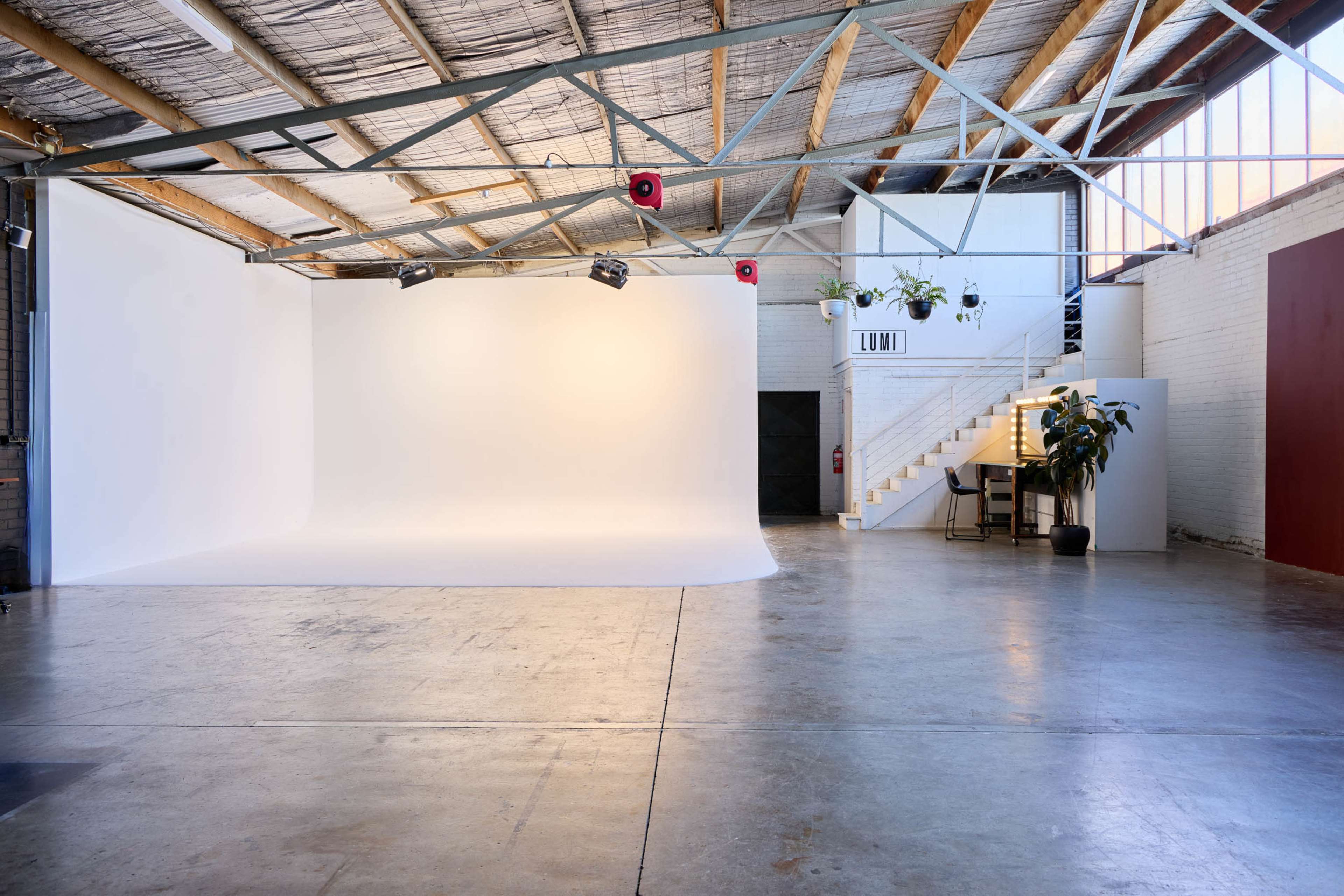 The image shows a spacious, well-lit studio featuring a large white backdrop, industrial-style ceiling beams, and a staircase leading to an upper area with a small workspace.