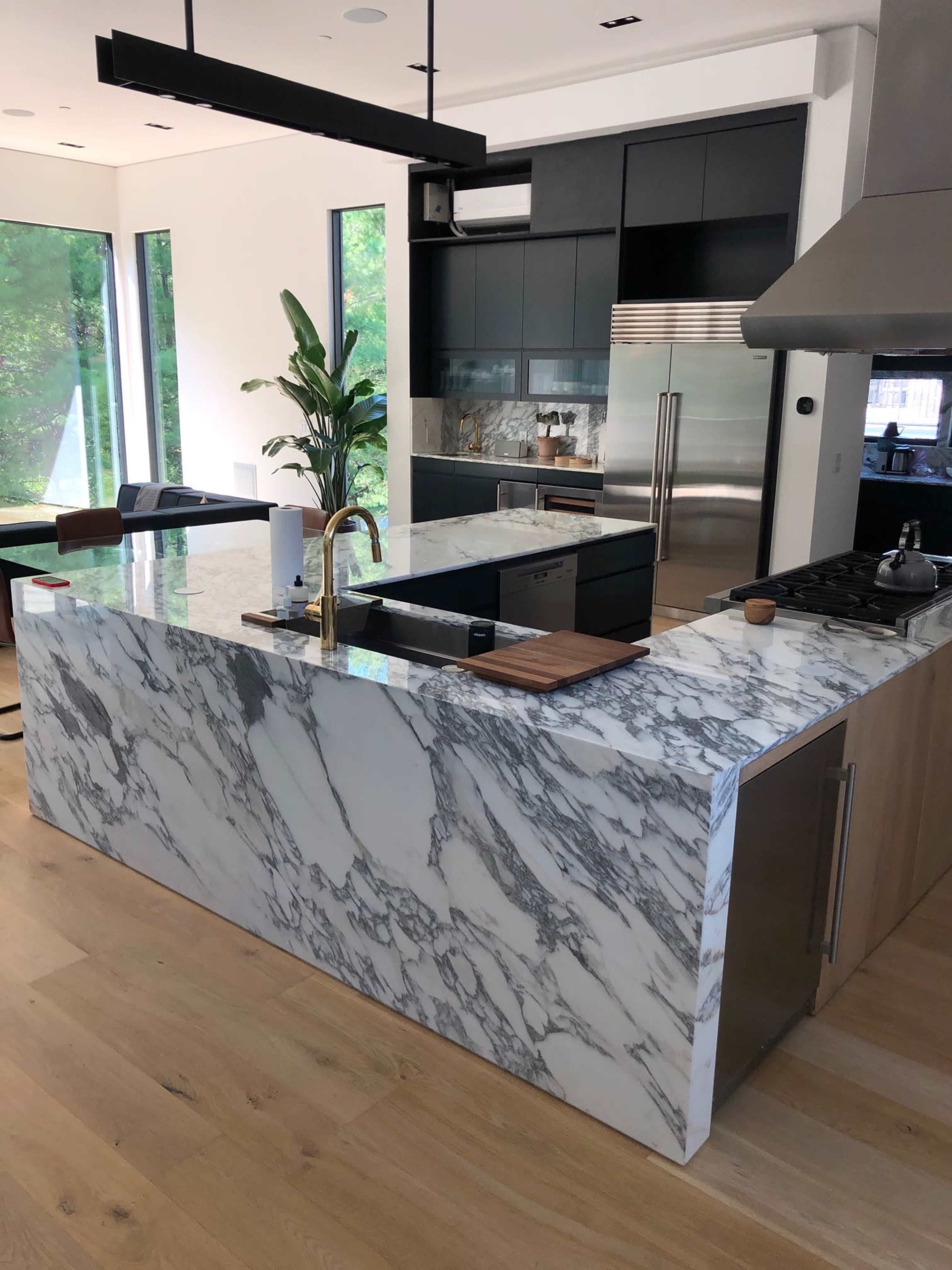 A modern kitchen with a large marble island, a stainless steel stove, and floor-to-ceiling windows featuring a view of greenery outside.
