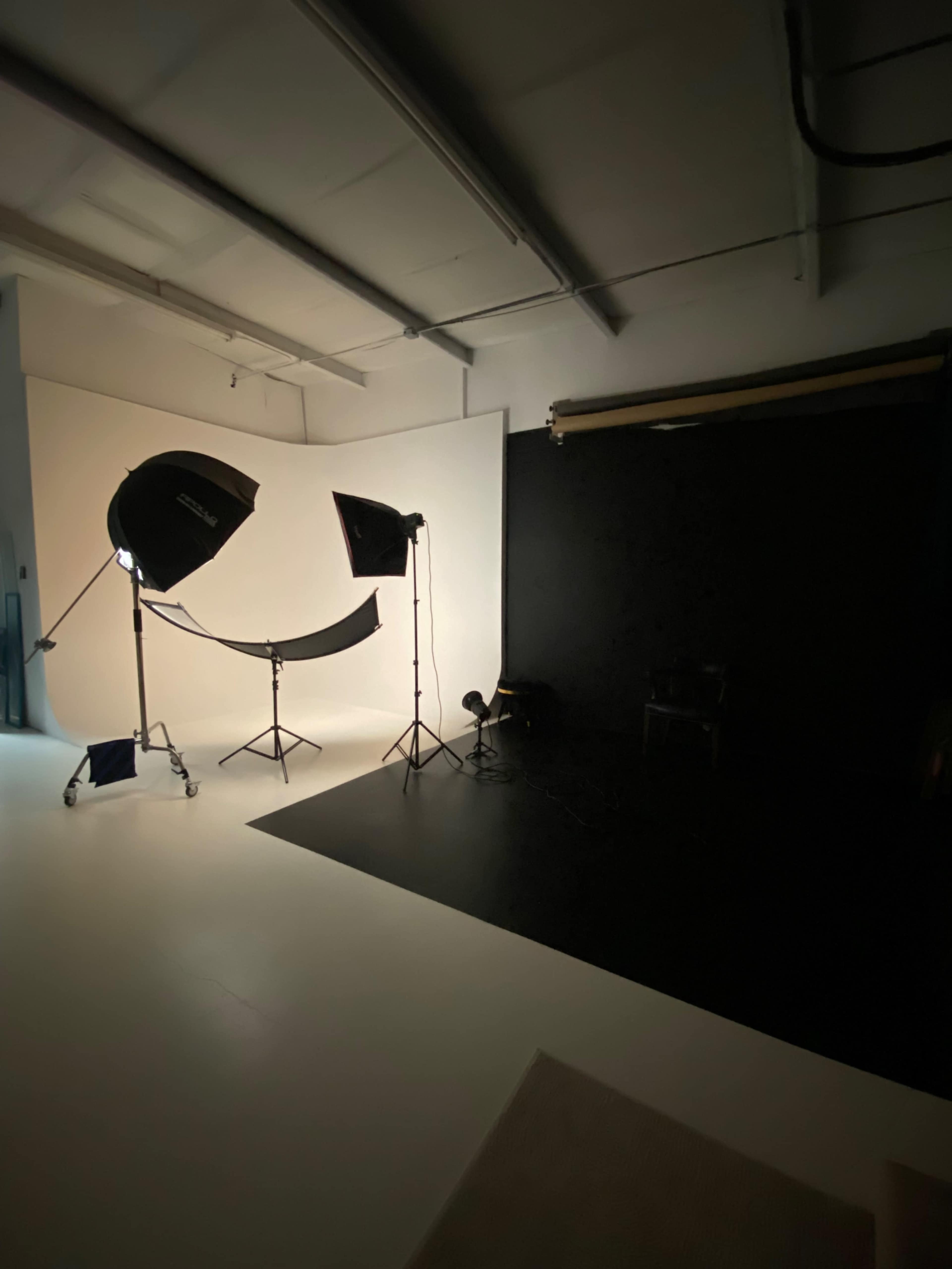 The image shows a photography studio with lighting equipment set up against a backdrop of white and black surfaces.