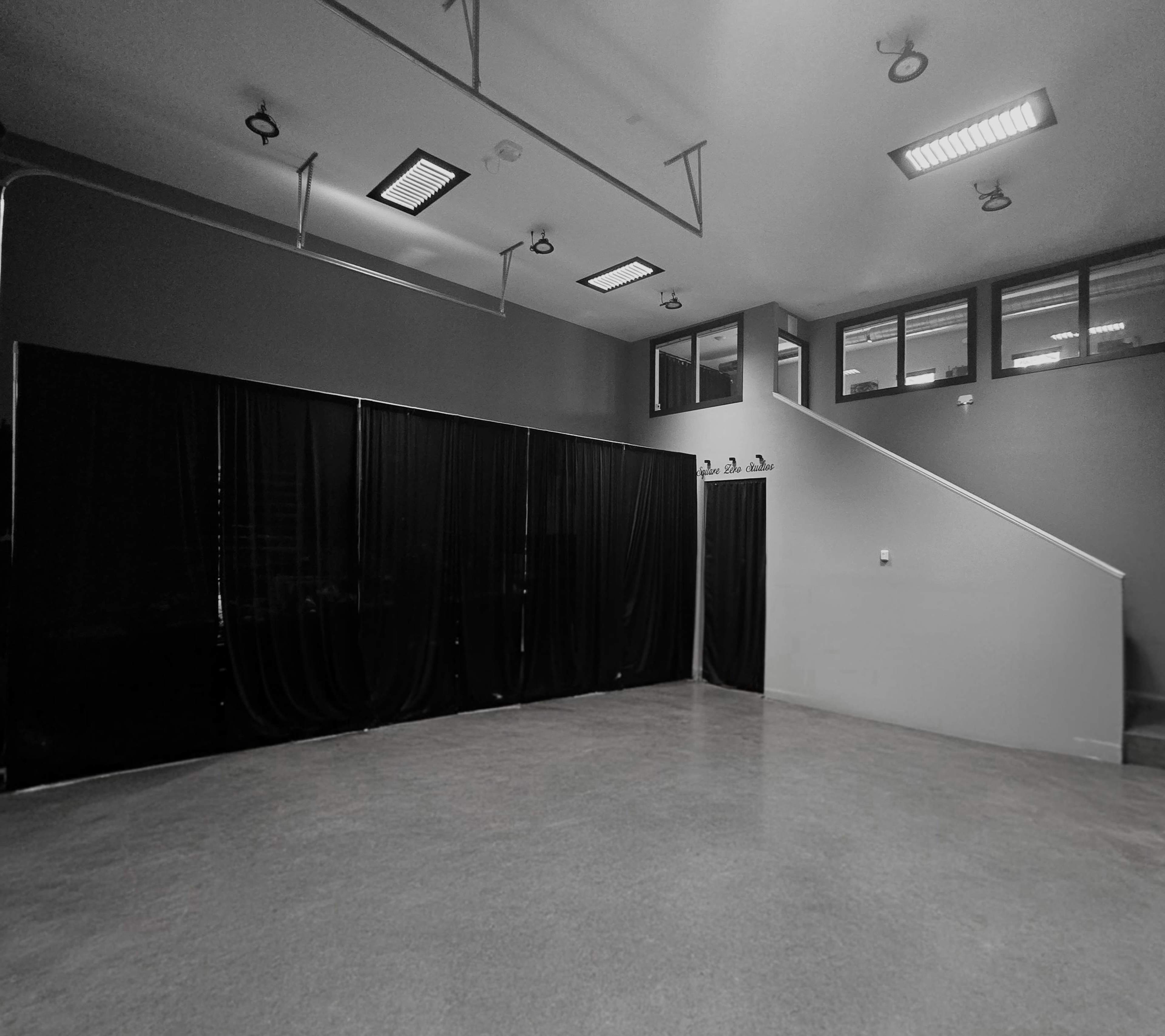 The image shows a spacious room with concrete flooring, black curtains on one side, and a staircase leading to a level with large windows.