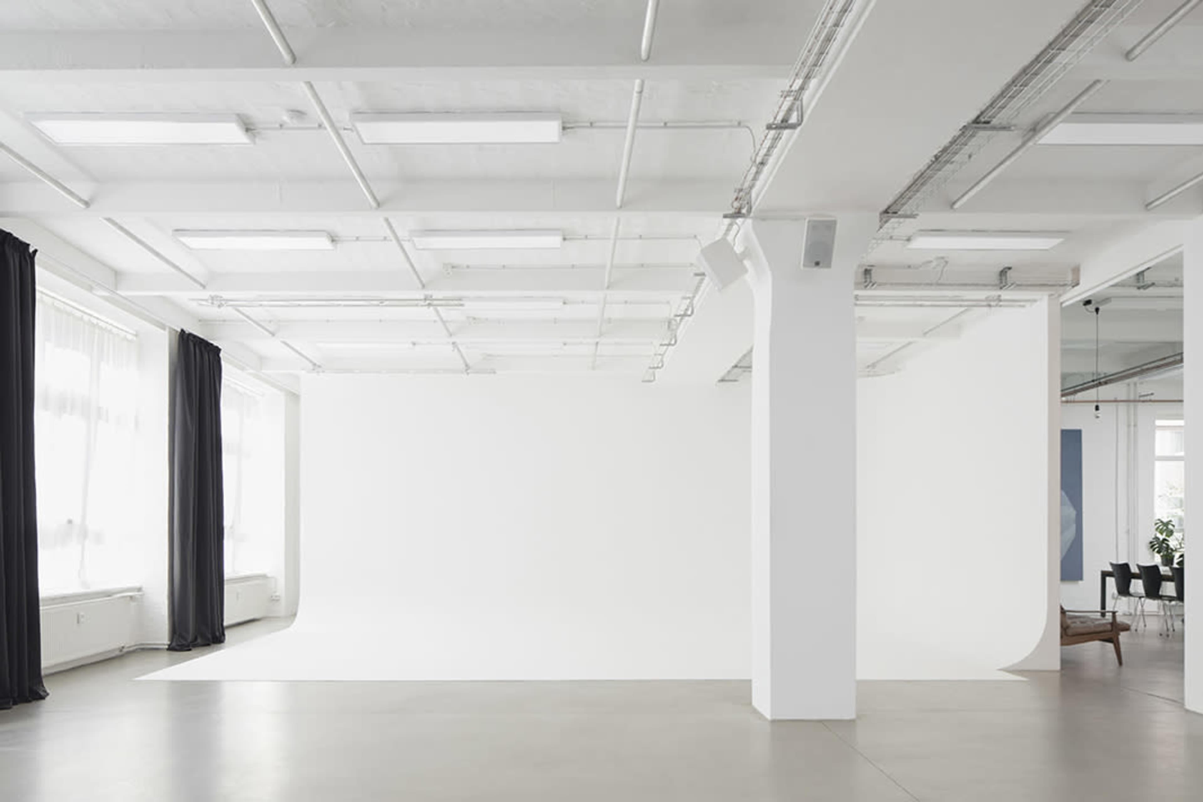 A spacious, empty studio with white walls and large windows, featuring a white backdrop and minimal furniture.