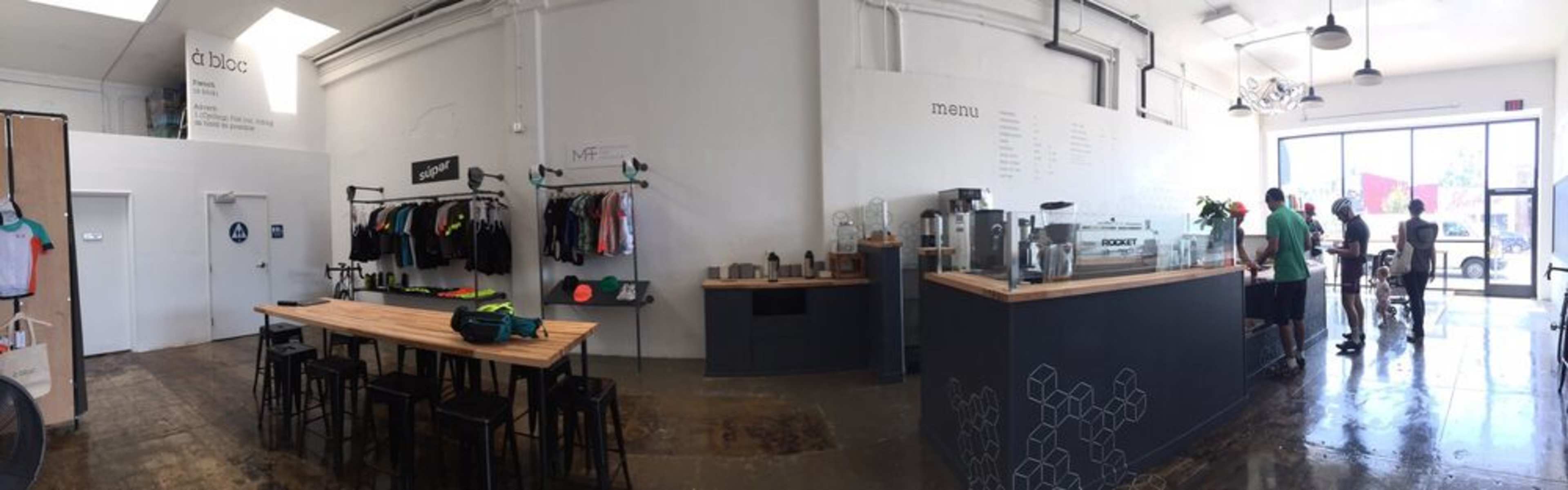 A spacious, modern retail space with a coffee area, a display of clothing on hangers, and a wooden counter with barstools where customers are interacting.