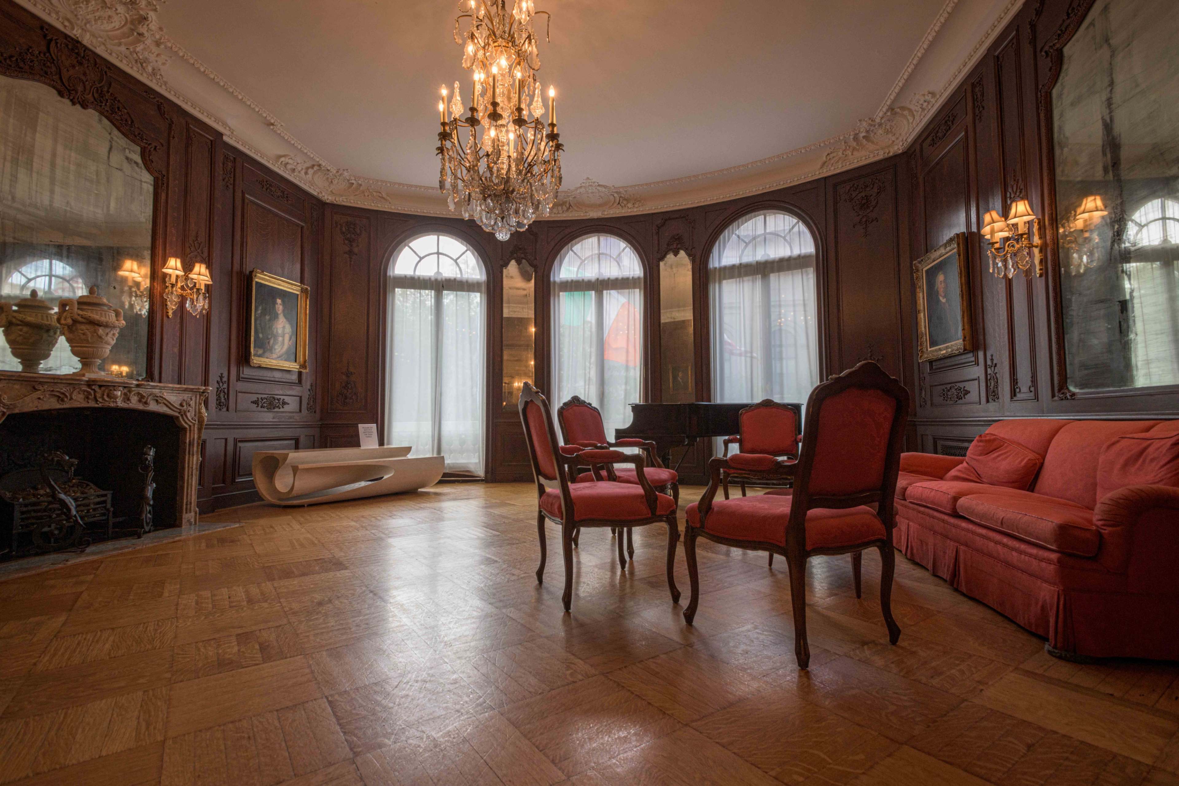 A spacious room with wooden paneling, large arched windows, a chandelier, and vintage furniture, including a sofa and chairs arranged around a coffee table.