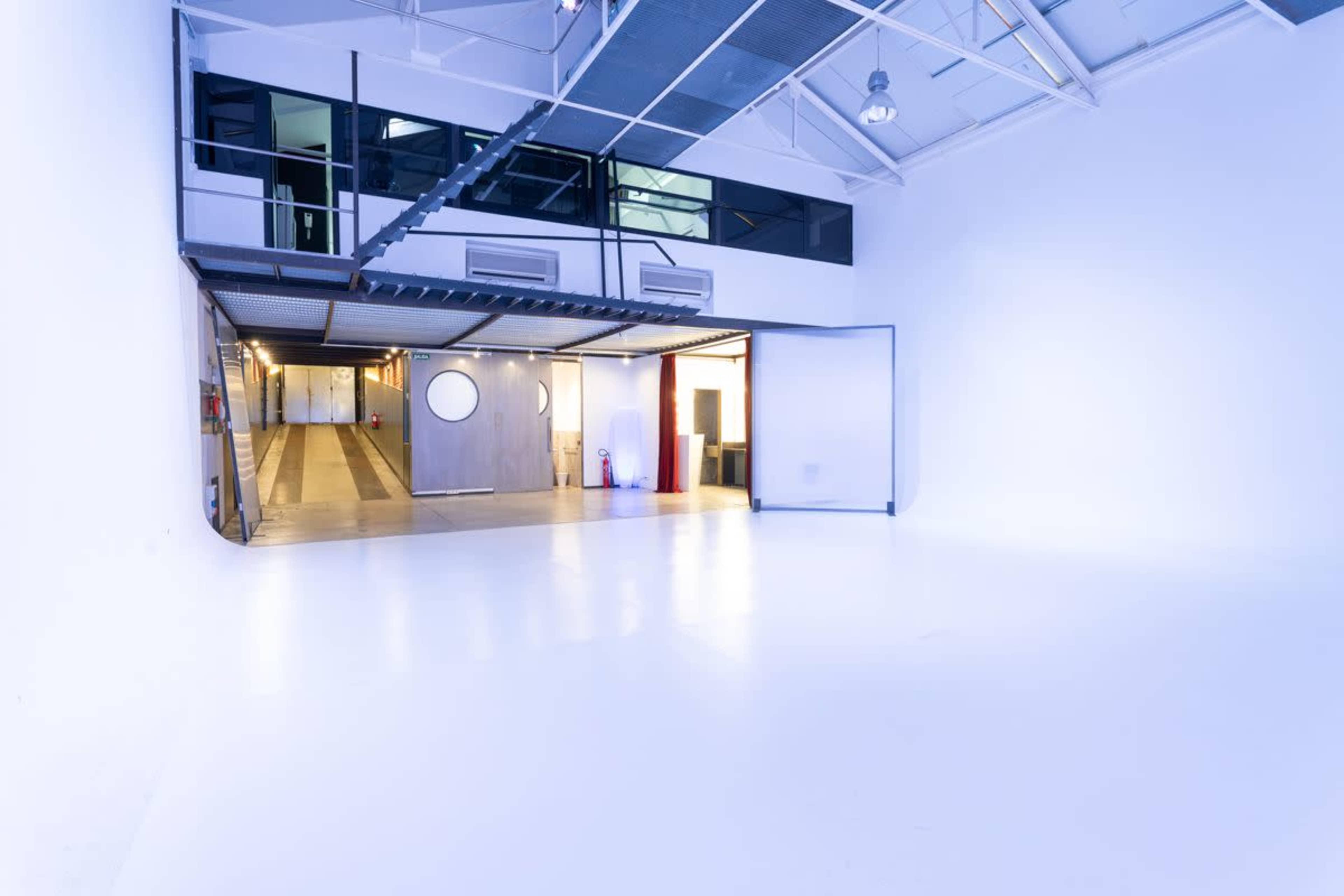The image shows a spacious, empty studio with white walls, high ceilings, and a mezzanine level overlooking a large entrance.