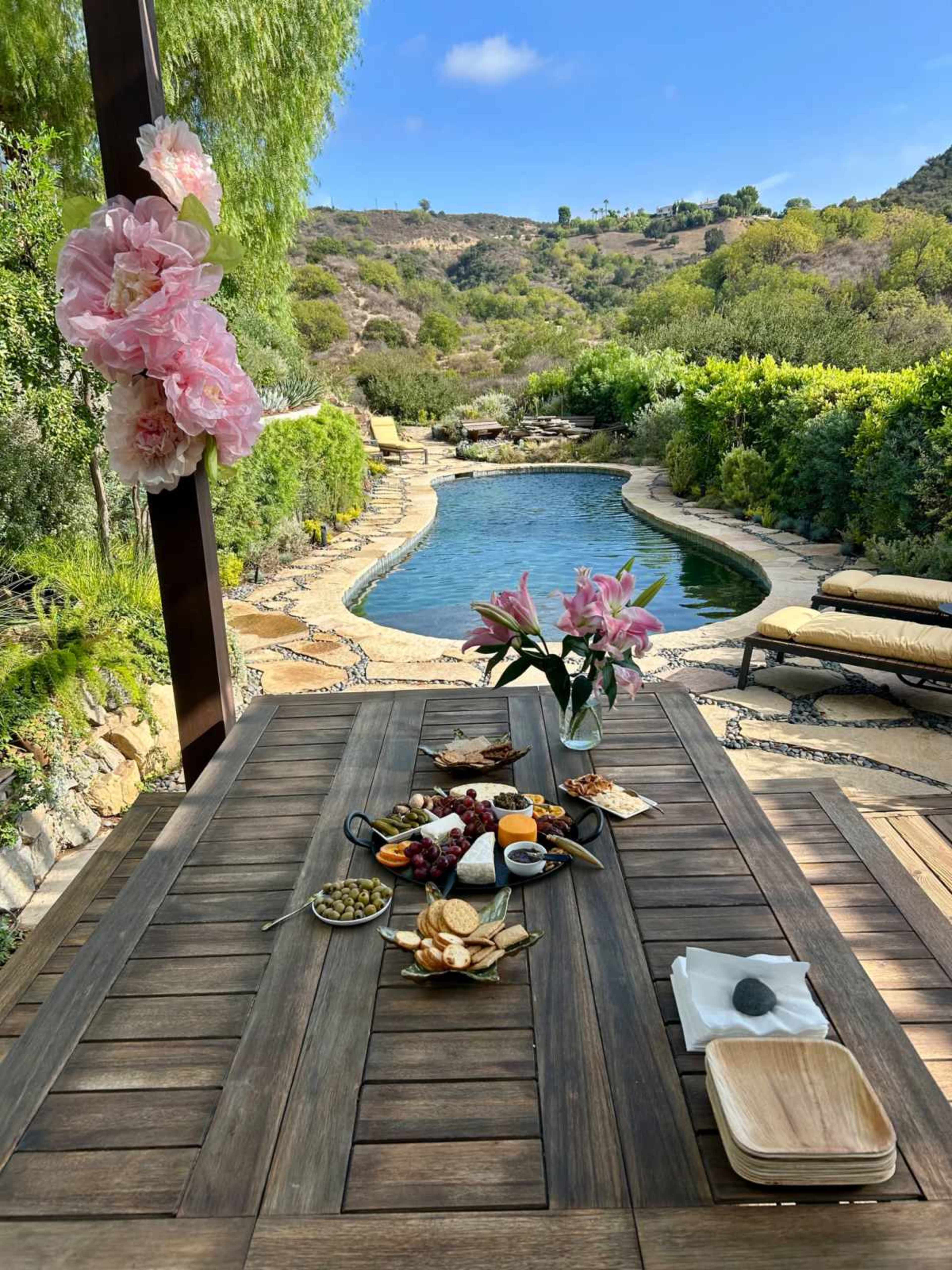 A table is set with various snacks and flowers, overlooking a serene pool and lush greenery under a clear blue sky.