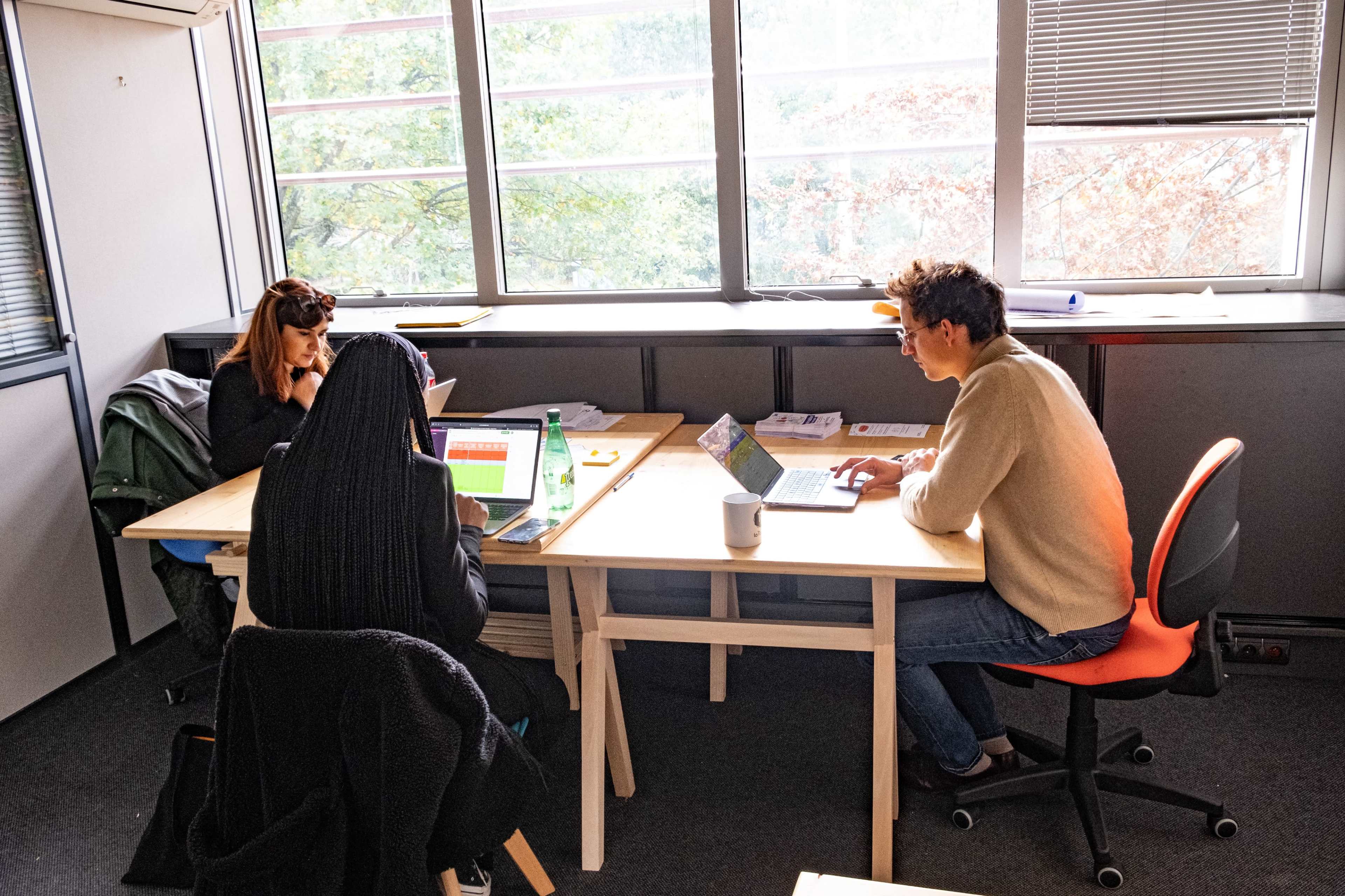 Three individuals are seated at a table working on laptops in a sunlit office space with large windows.