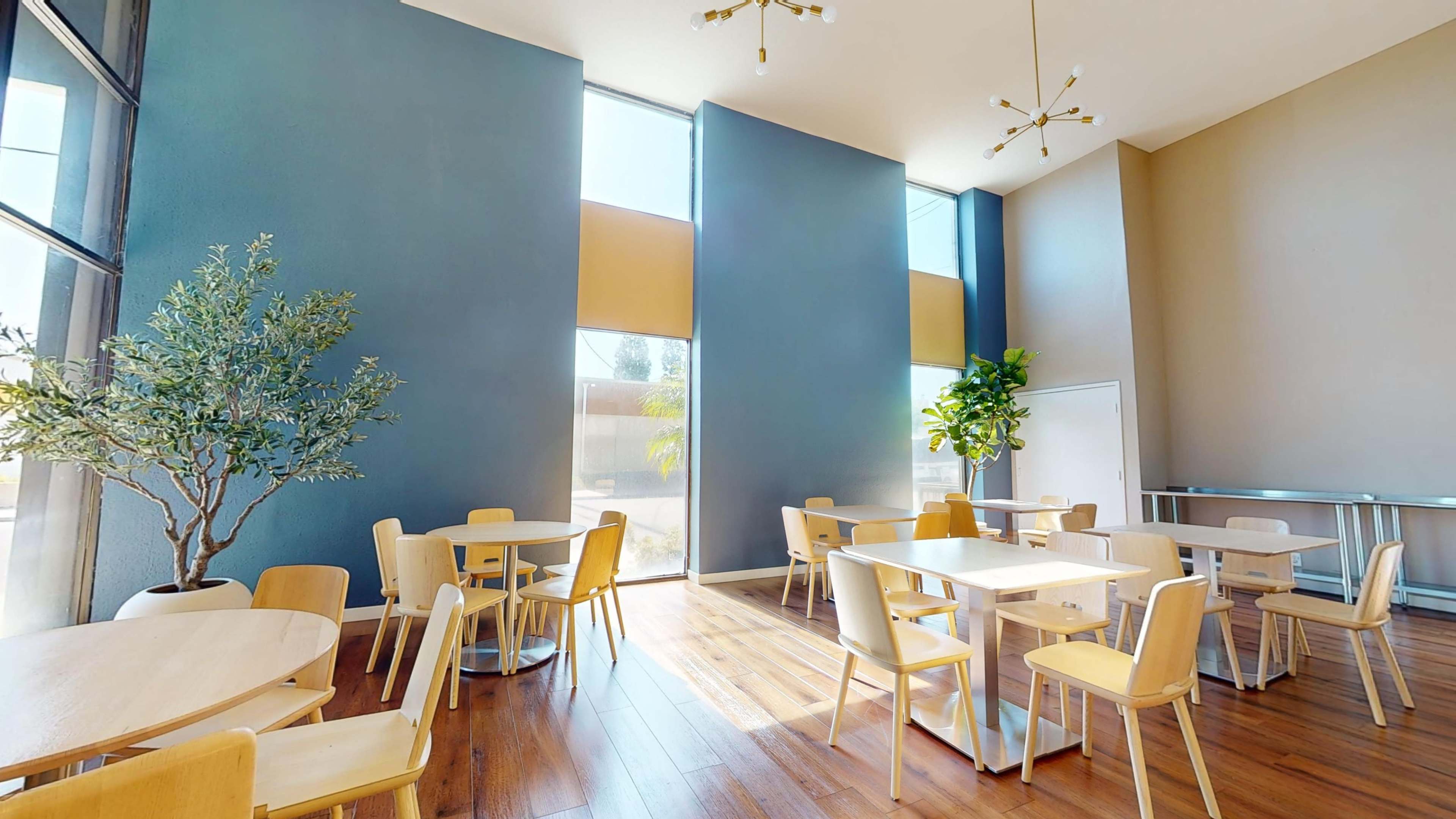 The image shows a spacious room with large windows, hardwood flooring, and several light wooden tables and chairs arranged throughout.