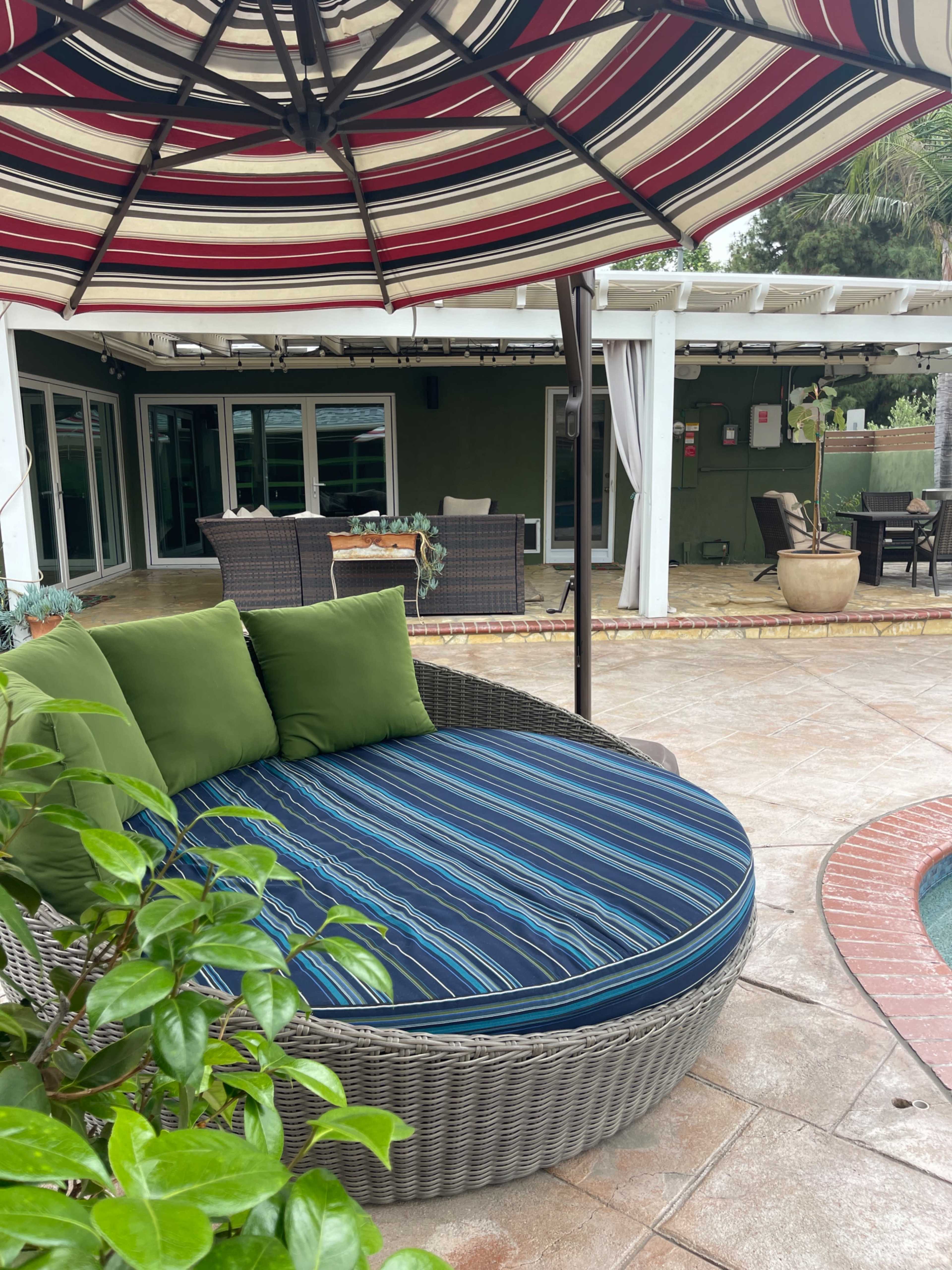 A round, striped outdoor lounge chair with green cushions is positioned under a red and white striped umbrella near a paved patio area.