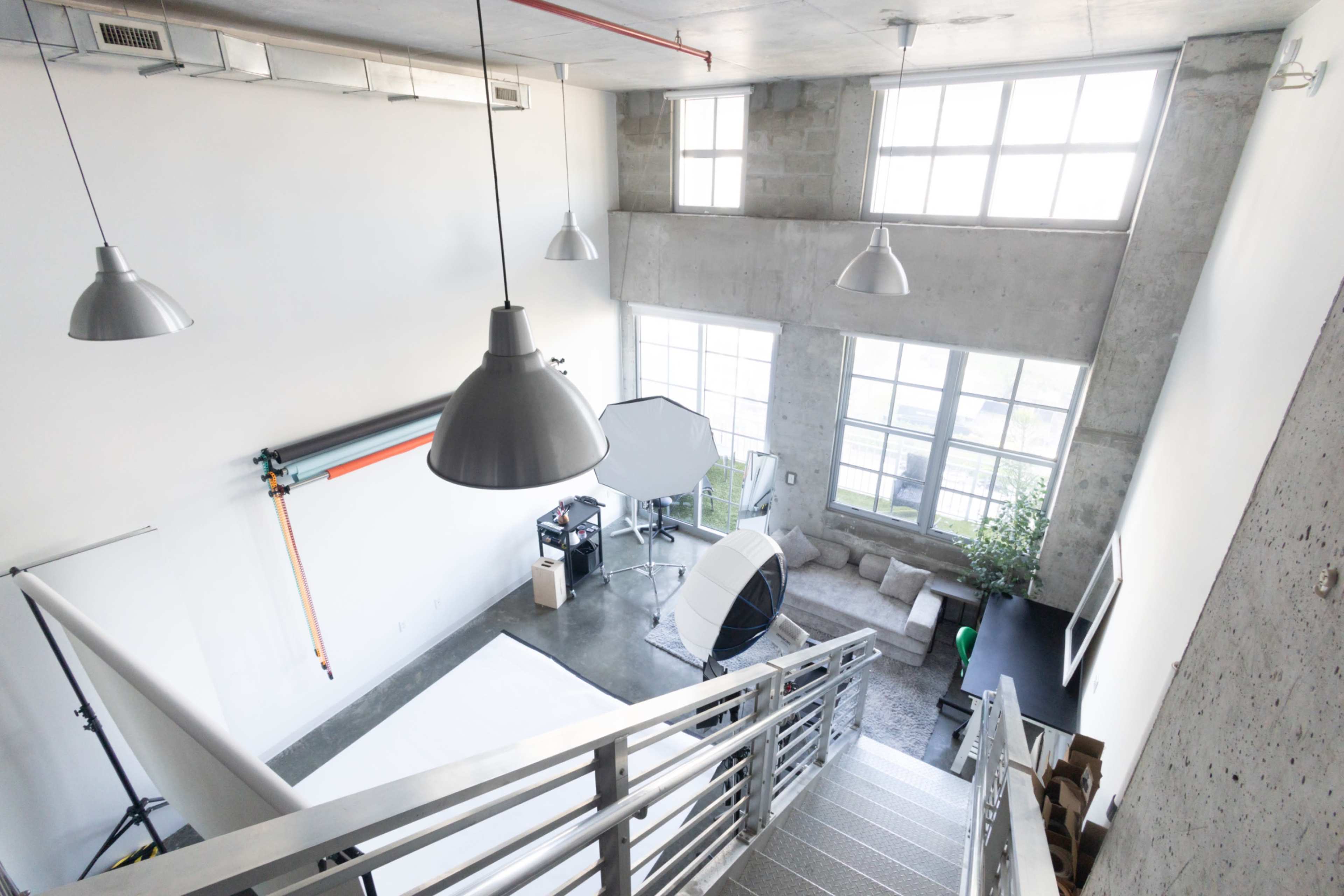 The image shows a well-lit photography studio featuring high ceilings, industrial-style concrete walls, and various photography equipment arranged around a large window.