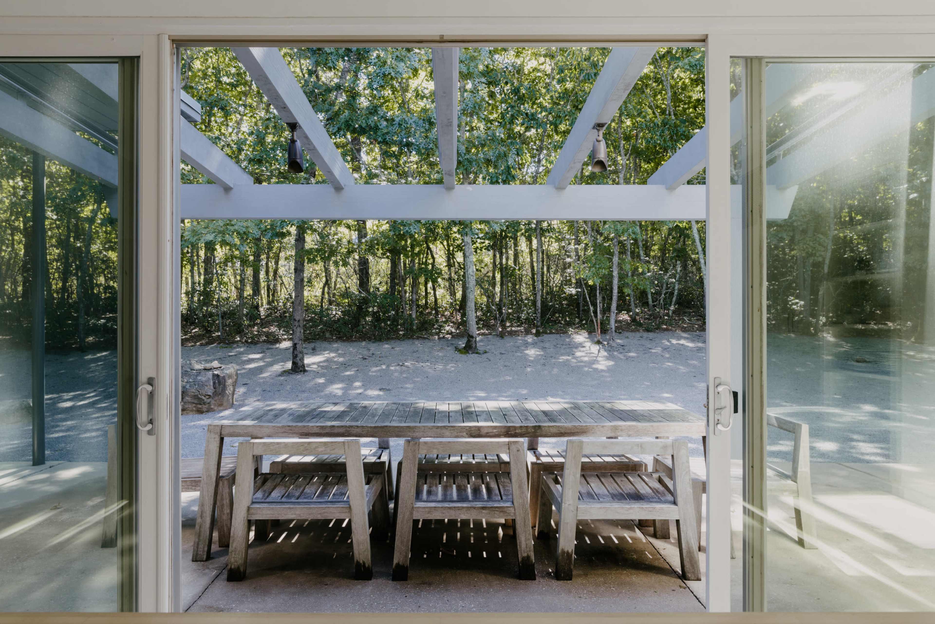 A wooden outdoor table and benches viewed through large glass doors, framed by a wooded area in the background.