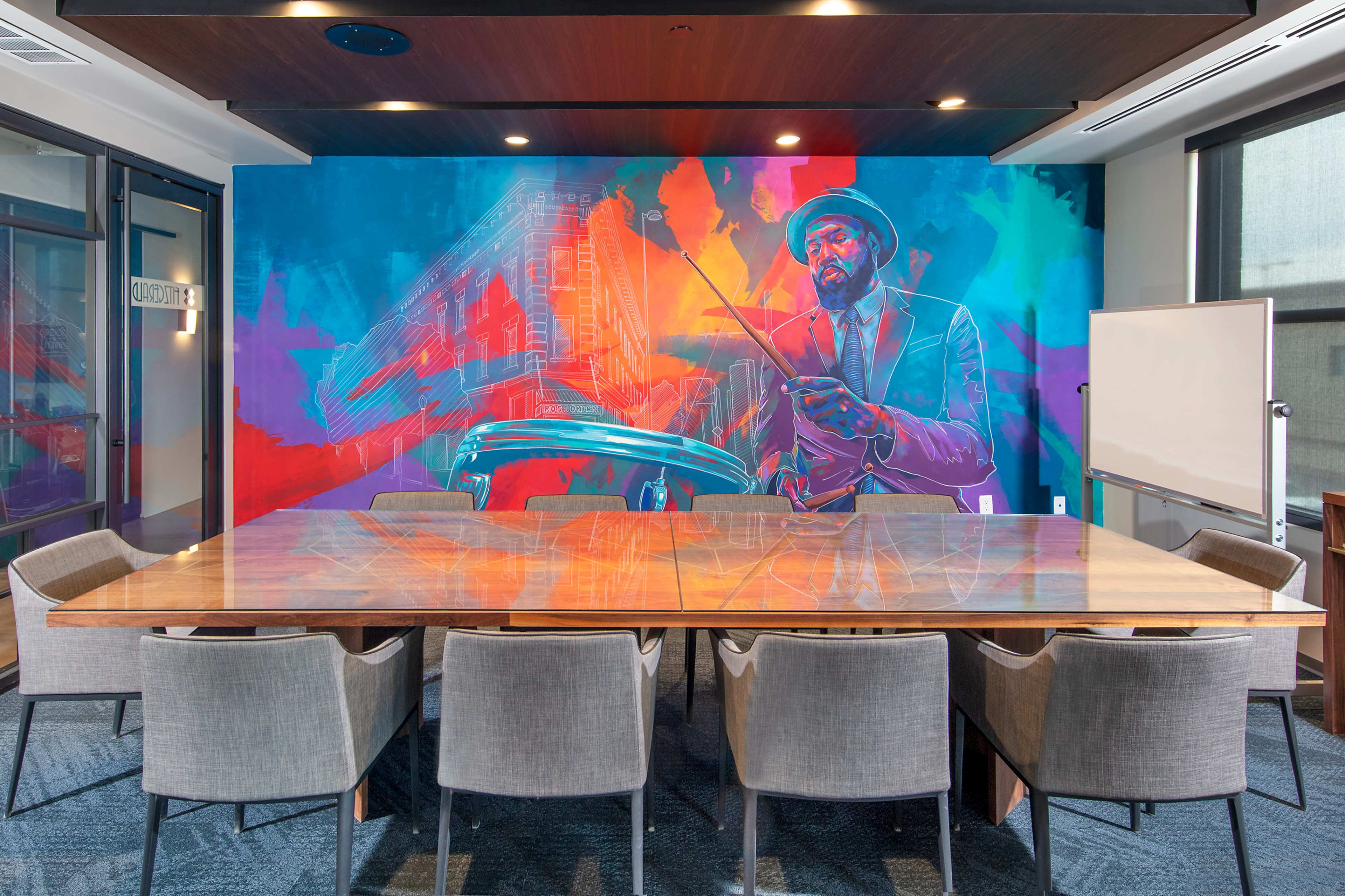 A modern conference room features a large wooden table surrounded by gray chairs, with a vibrant mural of a man holding a conductor's baton on the wall.