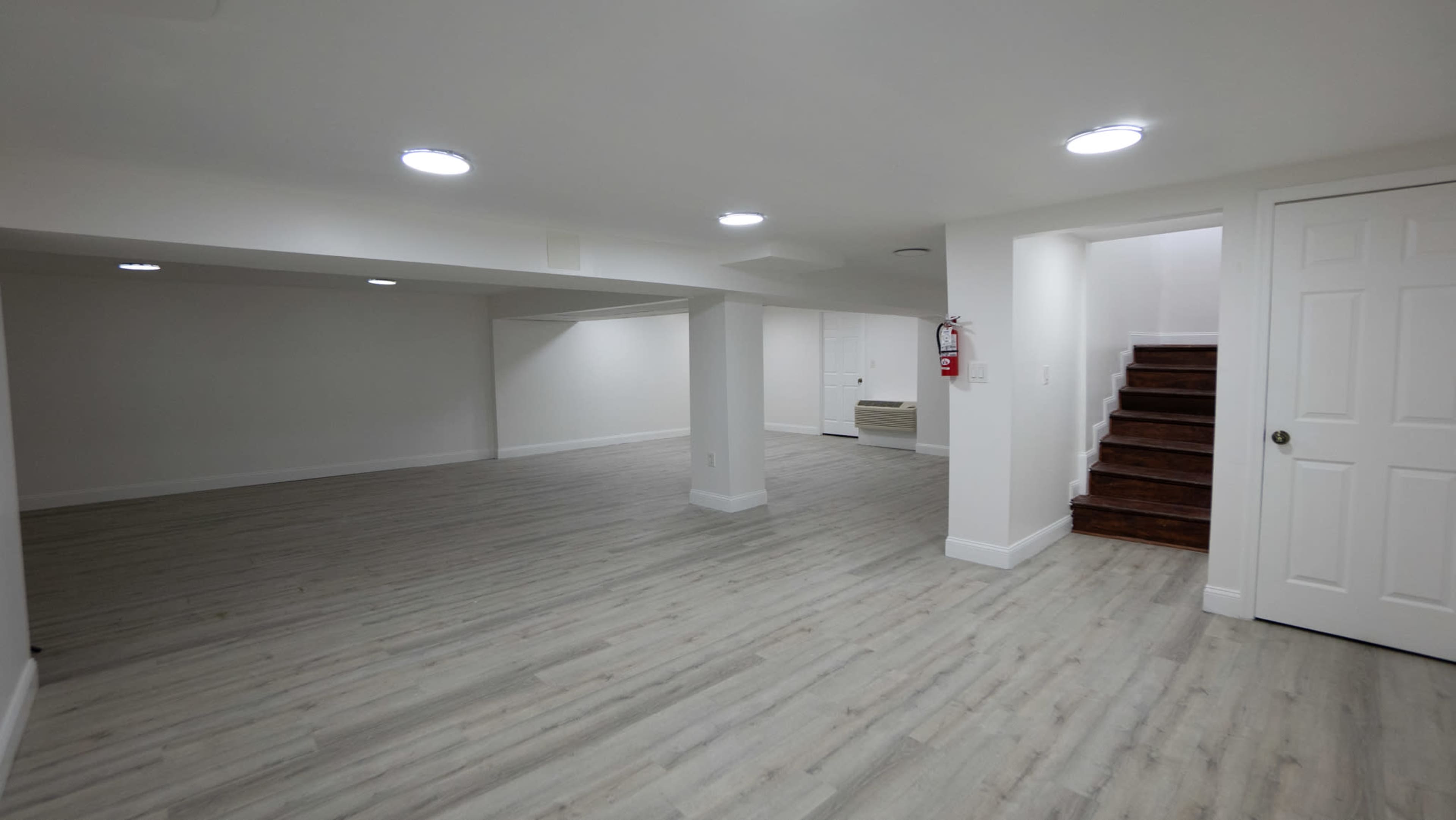 A spacious, empty basement area with light-colored flooring and a staircase leading to a door on the right.