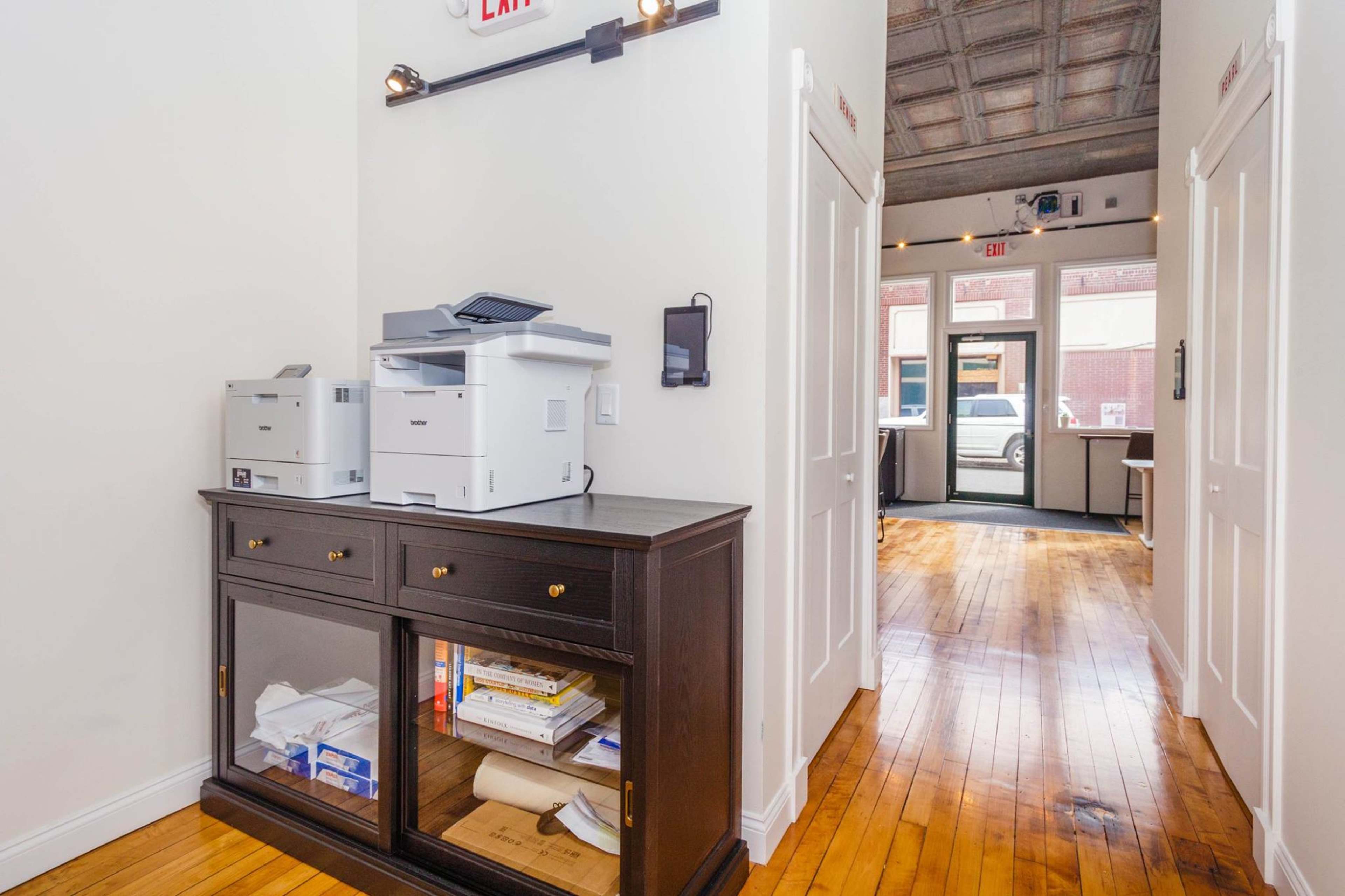 A hallway features a printer station with two printers on a cabinet and doors leading to an exterior area.