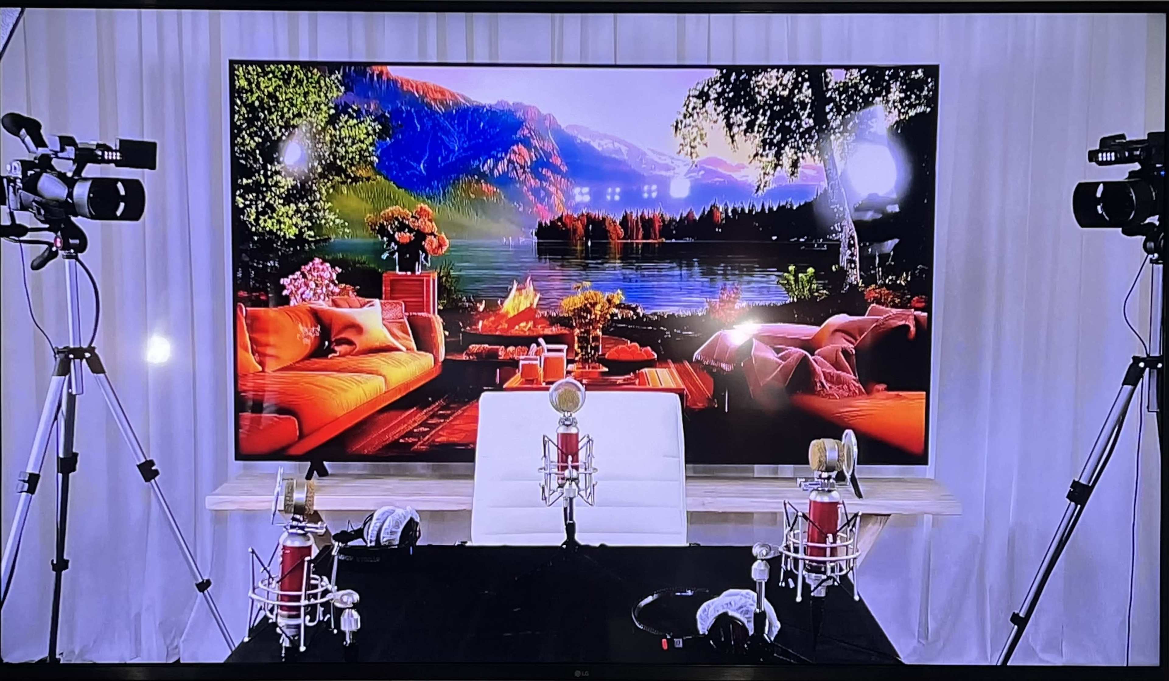 A large screen displaying a scenic landscape, flanked by two cameras and set up with microphones on a stage with a white backdrop.