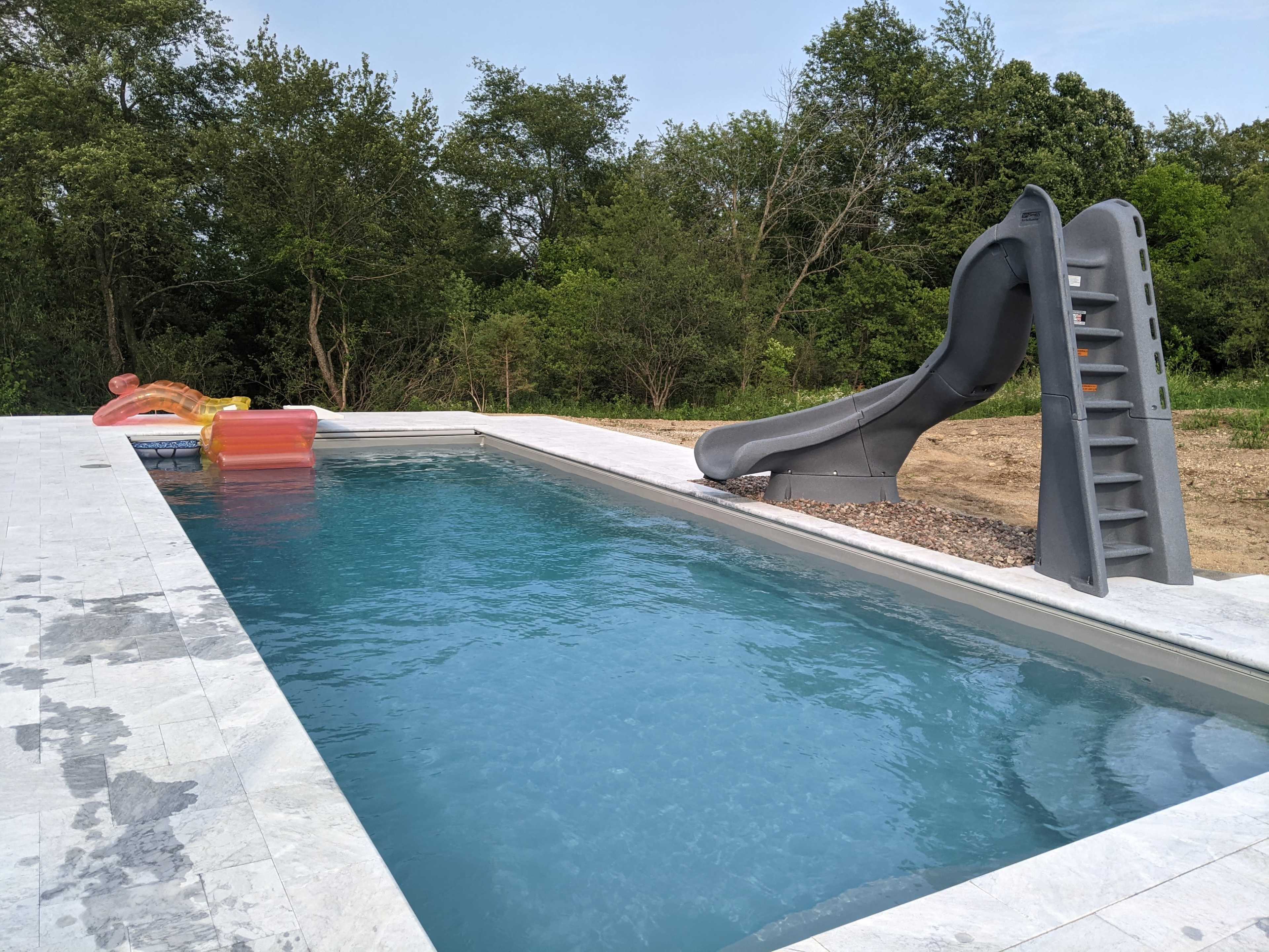 A rectangular swimming pool with a gray waterslide and colorful floating toys beside it, surrounded by greenery.