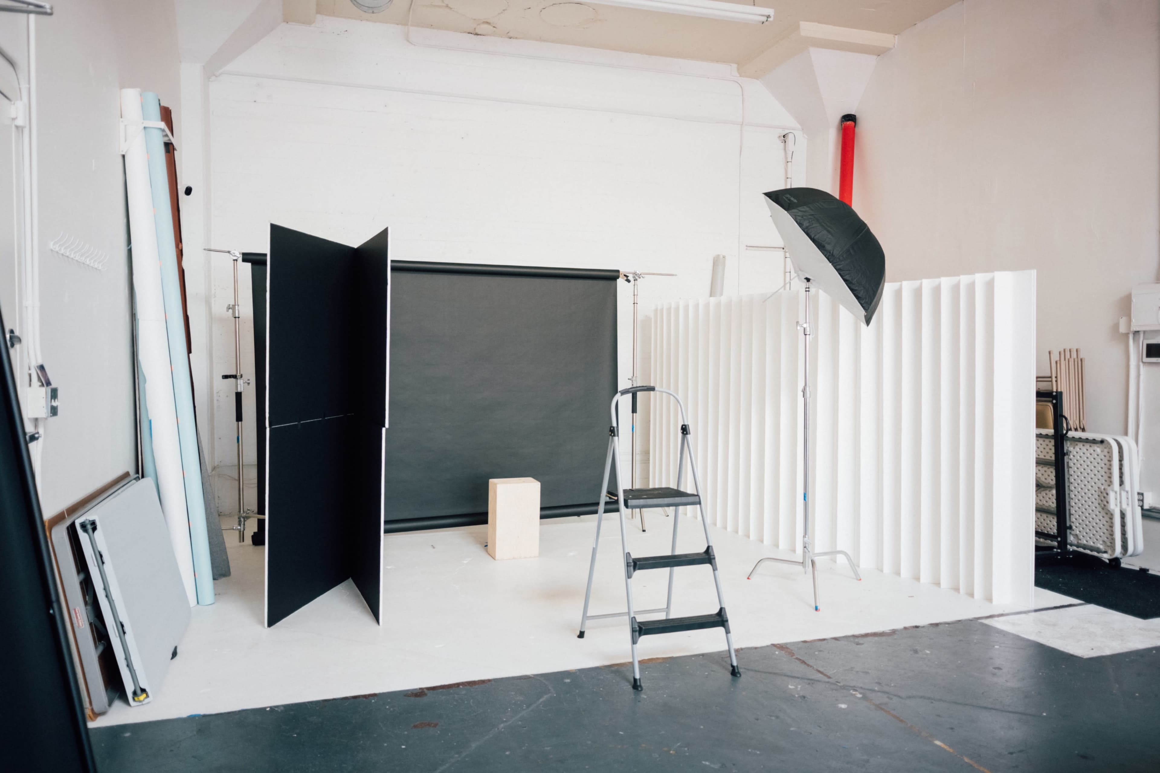 The image shows a photography studio setup with a black backdrop, two standing panels, a light umbrella, a ladder, and a wooden box.