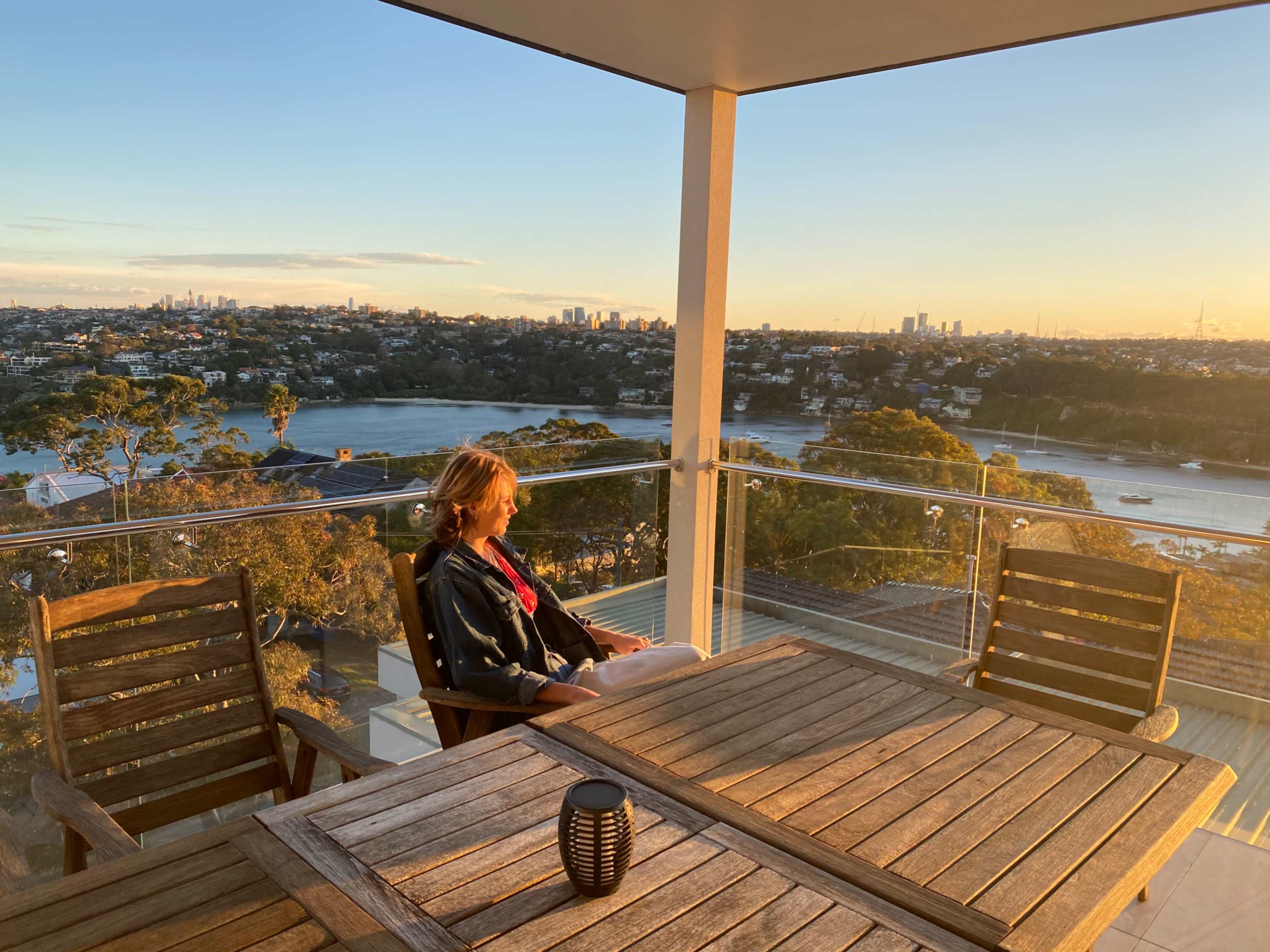 A woman sits on a balcony with a wooden table, overlooking a city skyline and river at sunset.