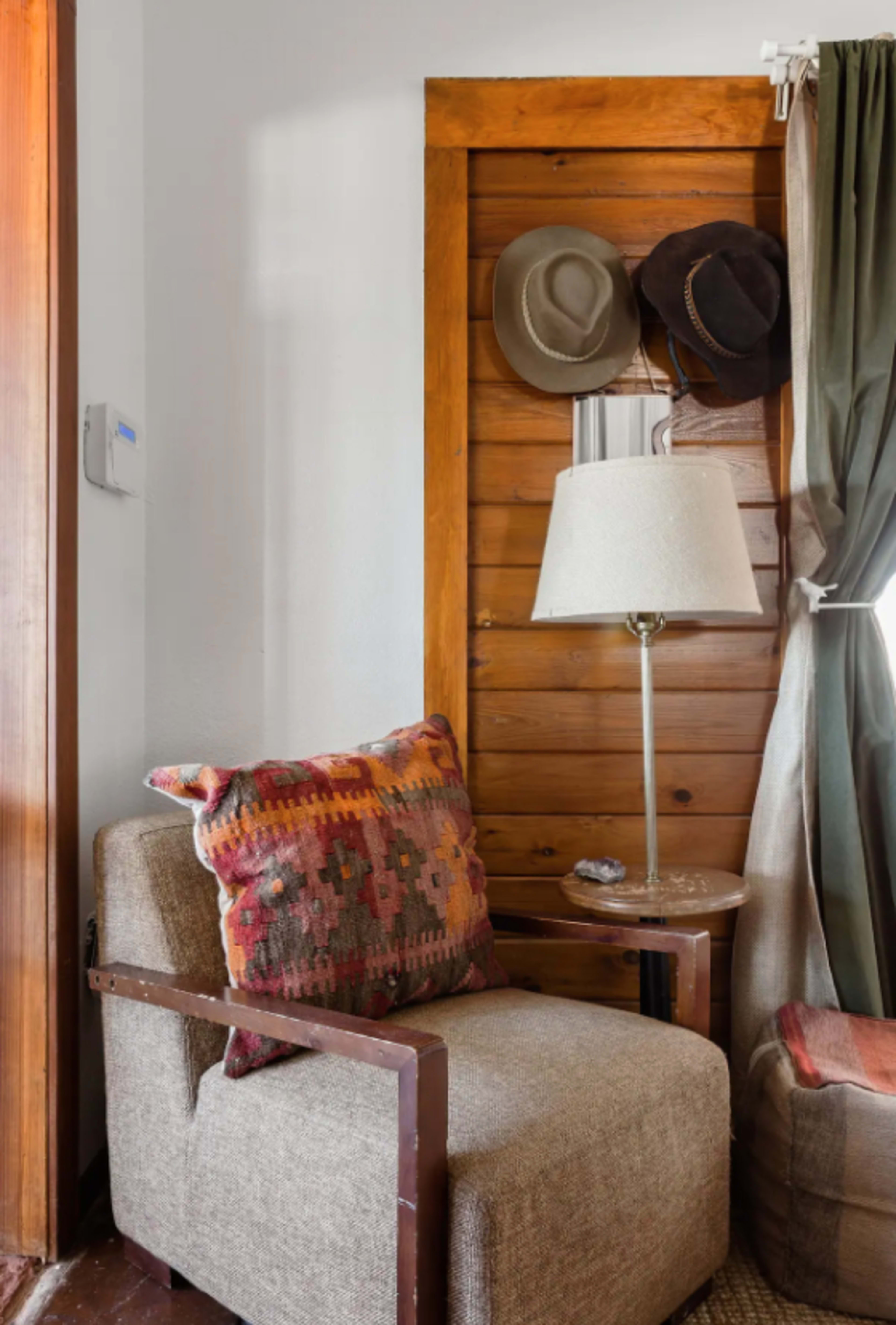 A cozy chair with a patterned pillow sits beside a small table and a lamp, adjacent to a wooden door with three hats hanging on it.