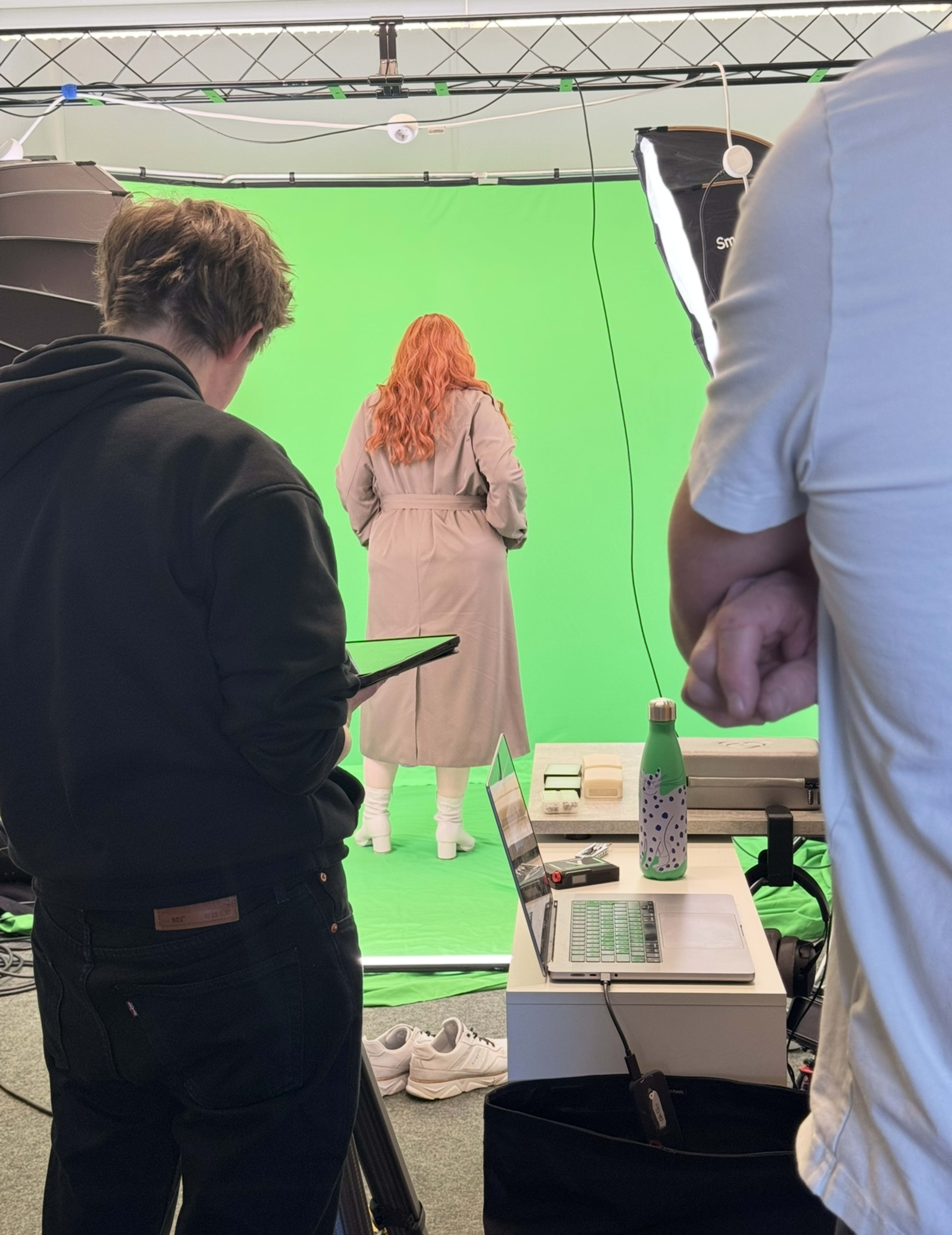 A person with long red hair wearing a beige coat stands in front of a green screen, while two individuals monitor a laptop and tablet nearby.