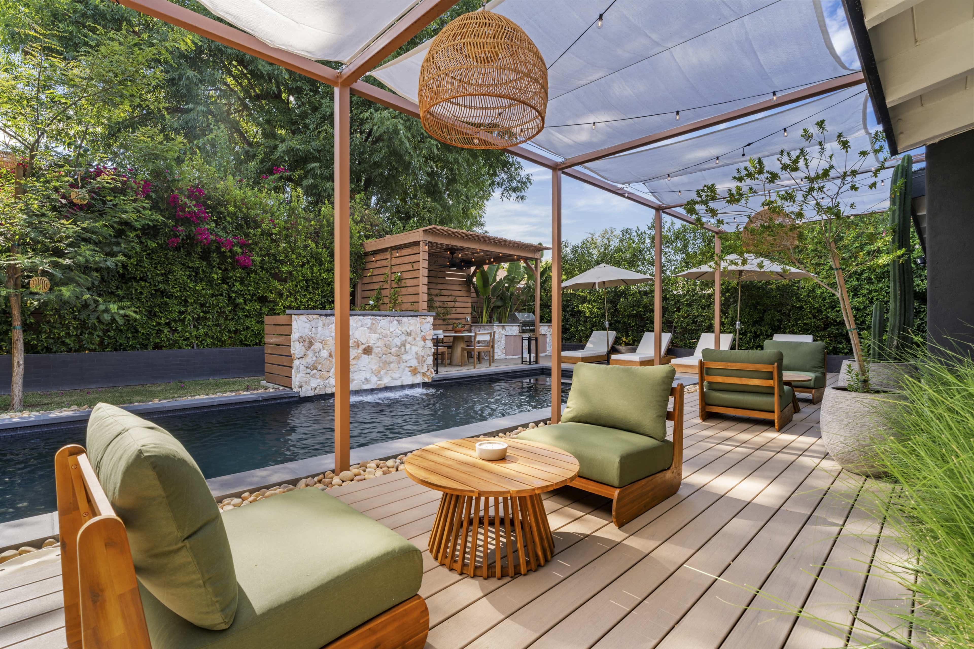 The image shows an outdoor terrace with two green lounge chairs, a wooden table, and a swimming pool, surrounded by plants and a bar area under a shaded structure.