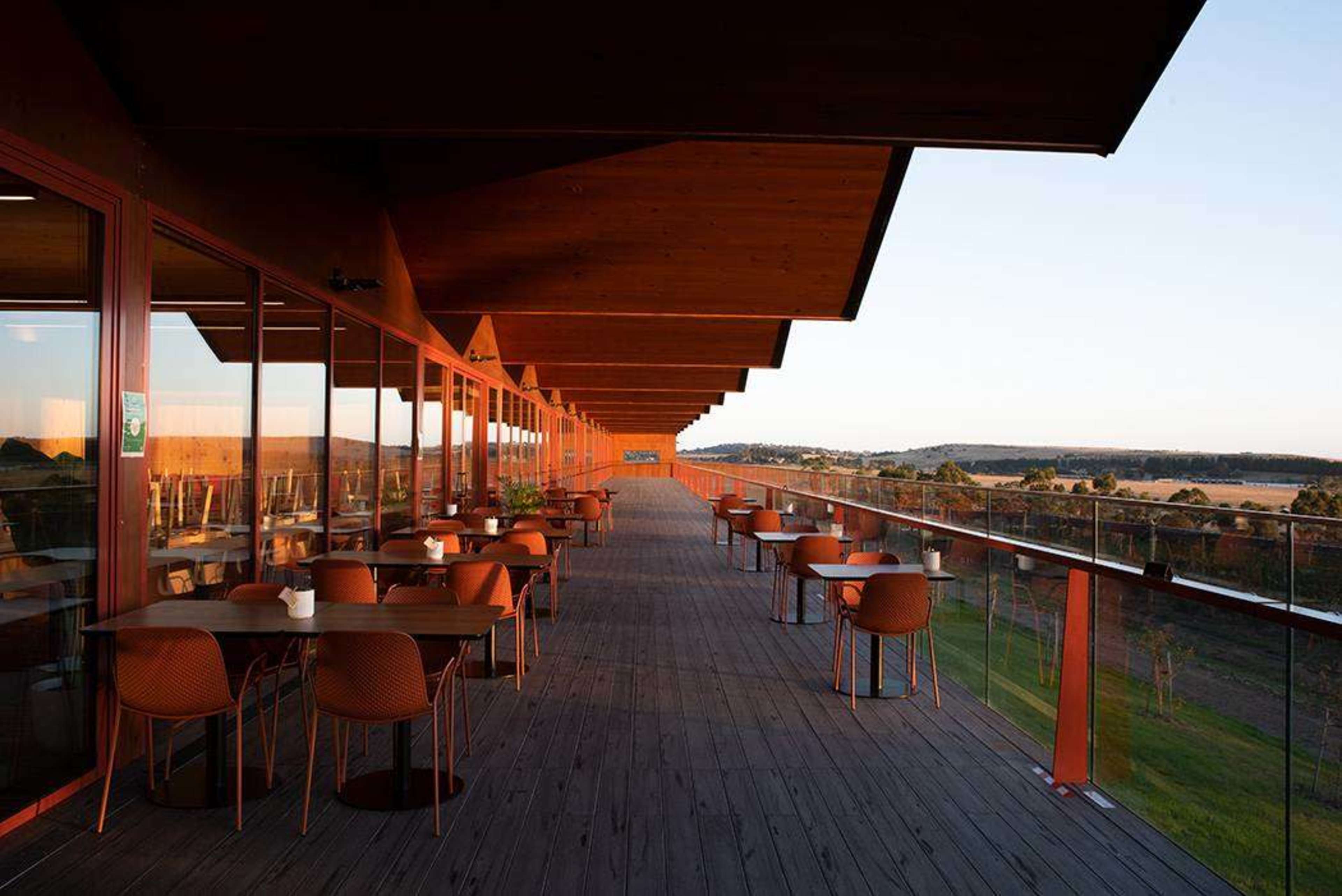The image shows a spacious outdoor restaurant terrace with orange chairs and tables, surrounded by greenery and a clear sky.