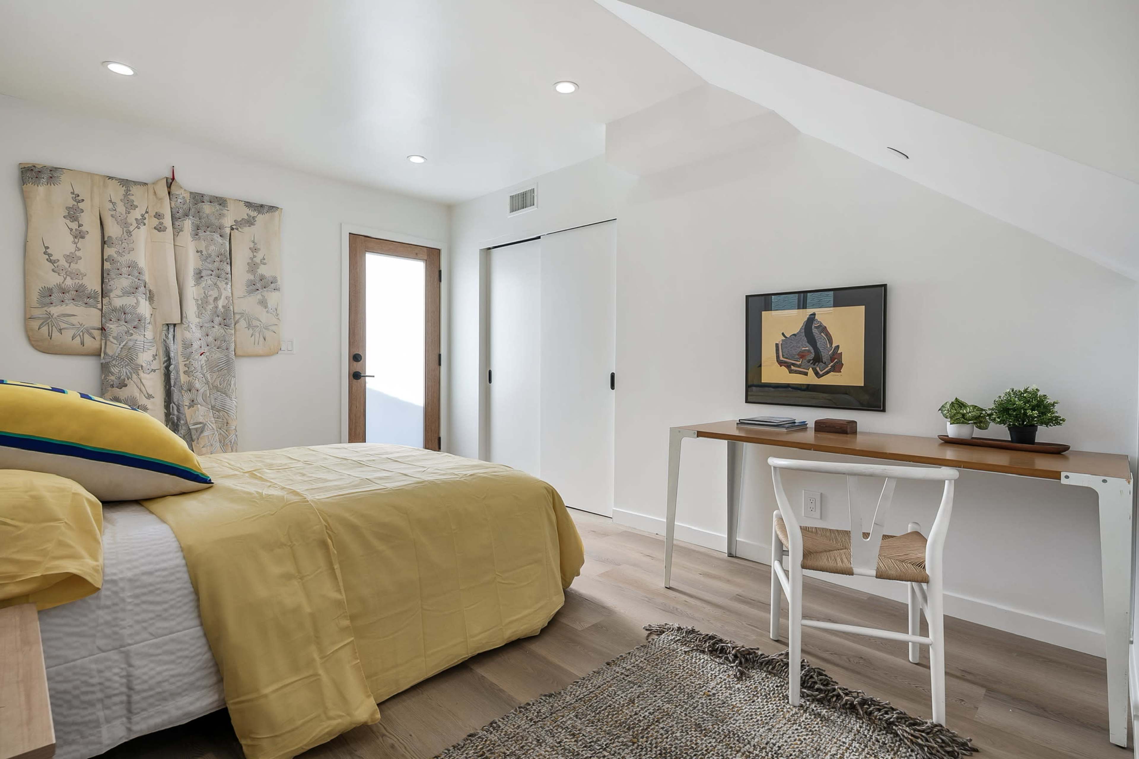 The image shows a bright bedroom featuring a yellow bedspread, a desk with a plant, and a door leading outside.