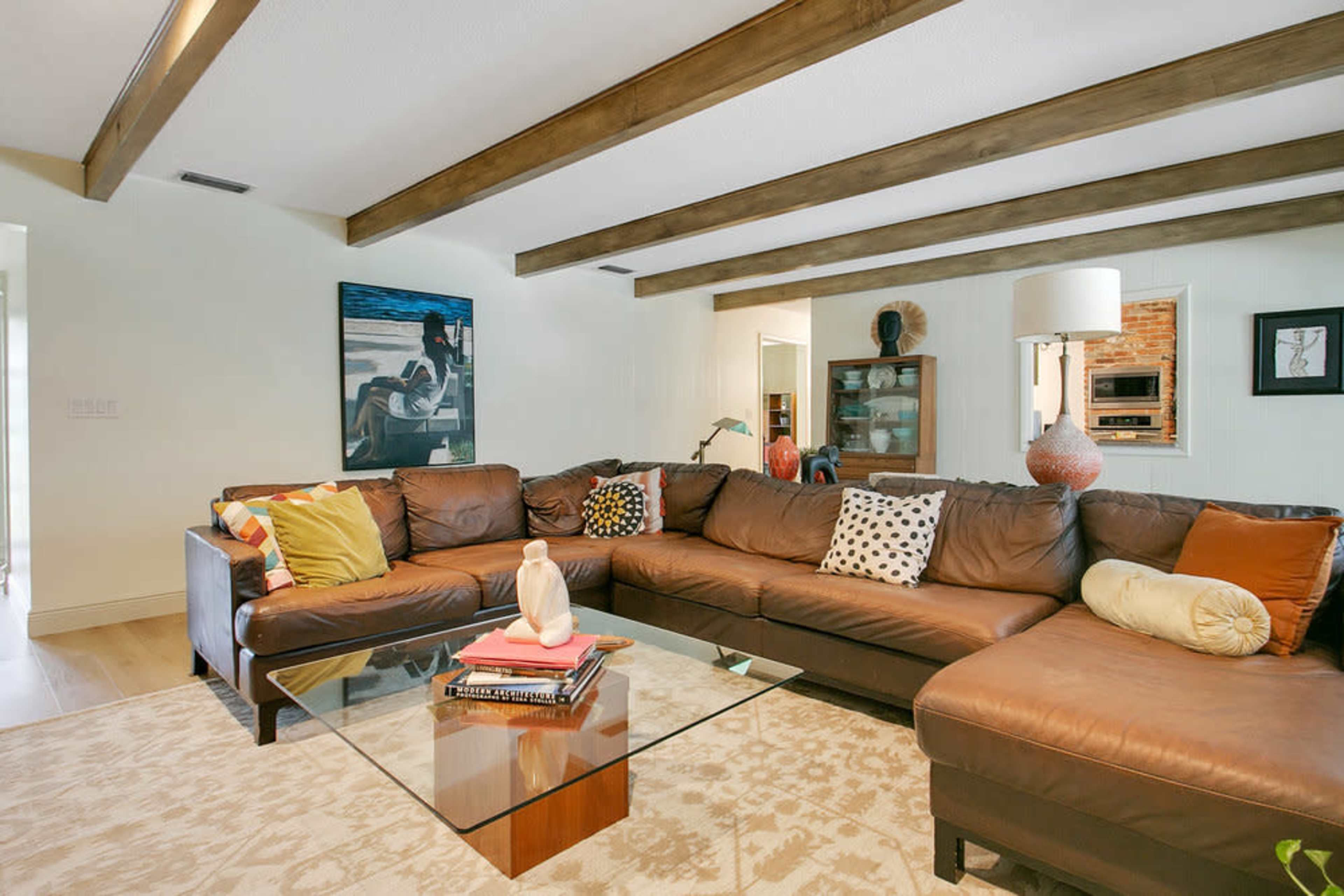The image shows a living room with a large brown leather sectional sofa surrounding a glass coffee table, accented by decorative pillows and a wall art piece.