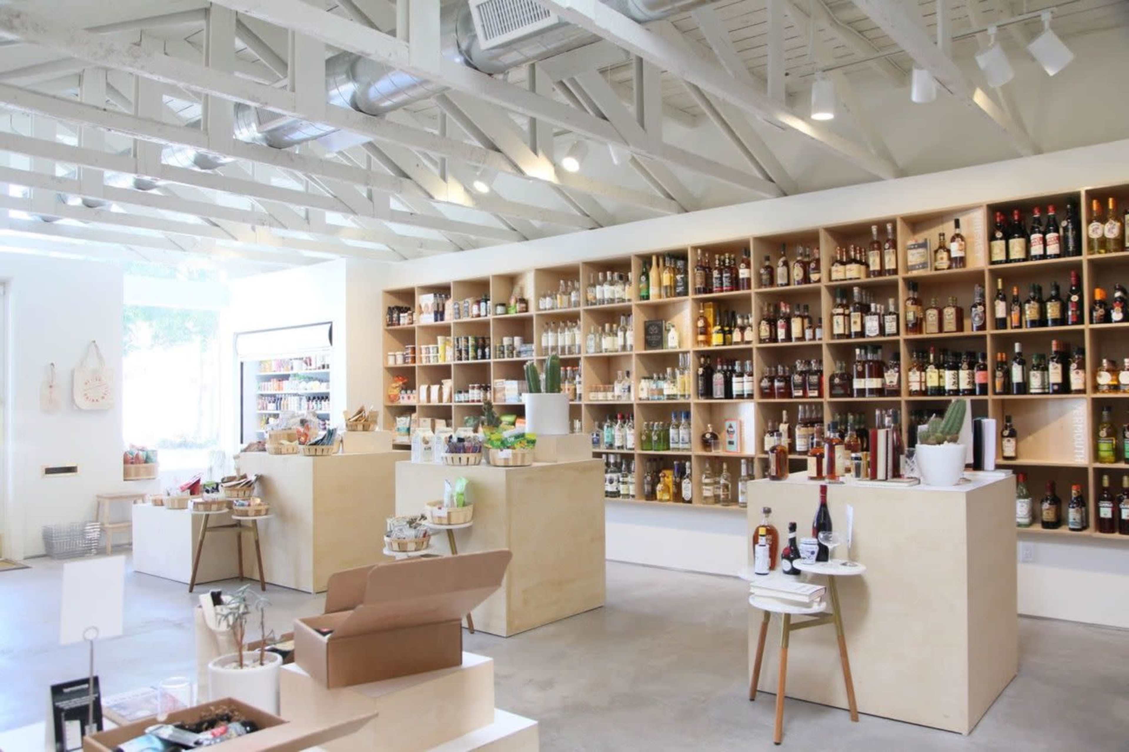 The image shows a modern retail space with wooden shelving displaying various bottles and products, along with minimalist furniture and bright lighting.