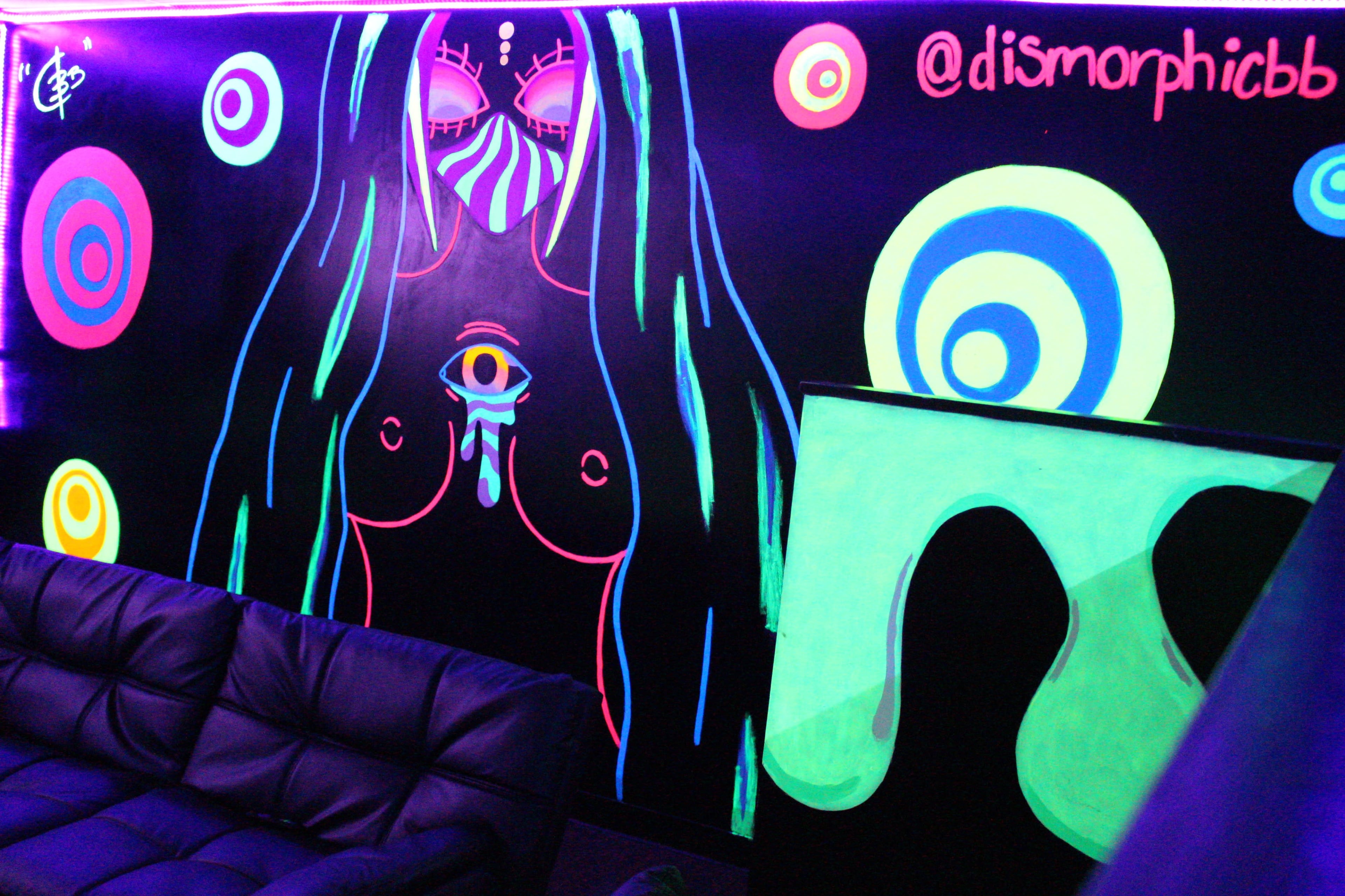 The image shows a dimly lit room with a neon-colored mural on the wall, featuring abstract shapes and a stylized figure with large eyes and an eye on the chest.